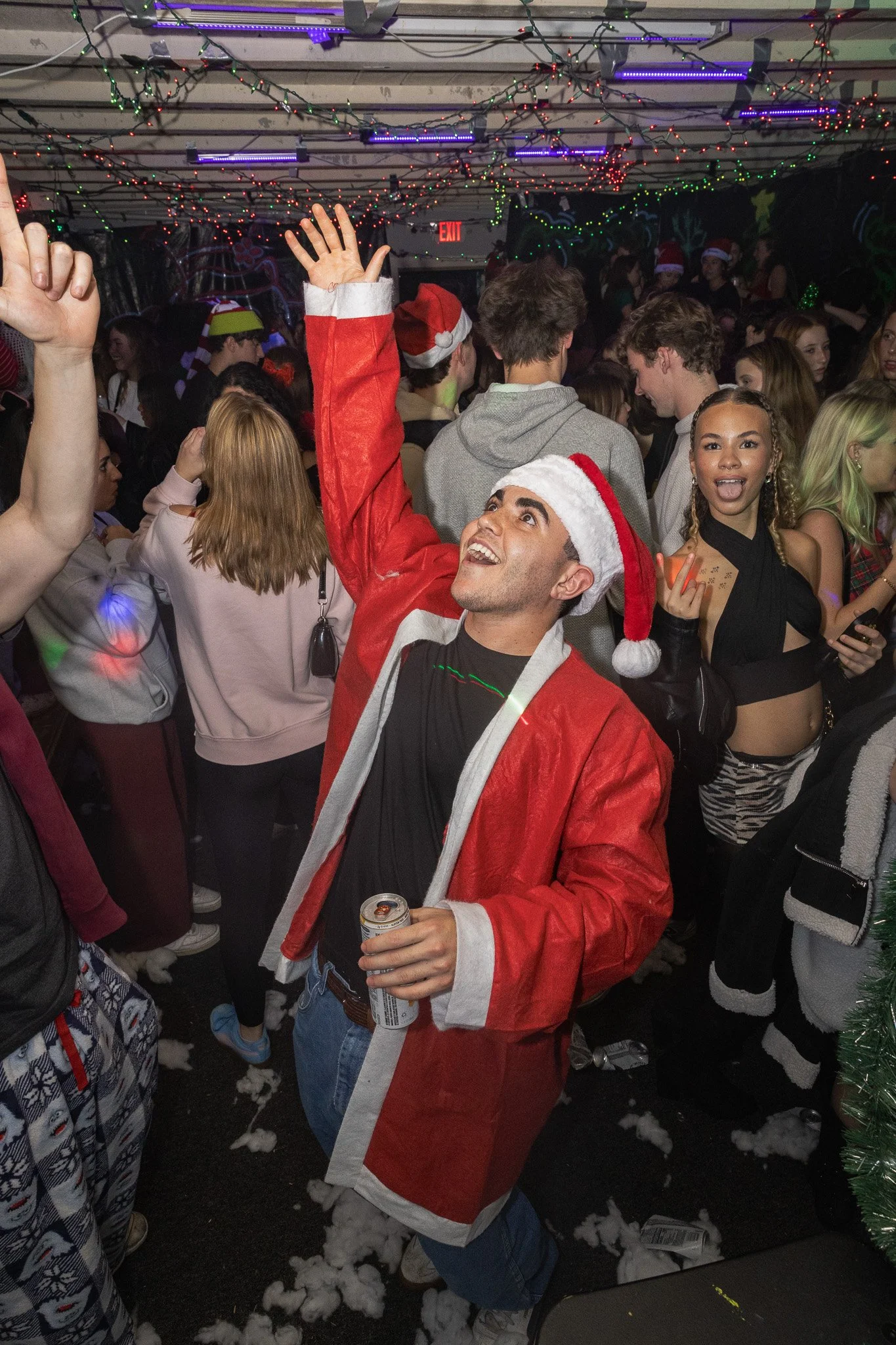 A group of young adults celebrating Christmas at a party with Christmas decorations and lights. One man in the foreground is wearing a Santa hat and a red Santa costume jacket, holding a drink, and raising his hand in the air. The background shows ot