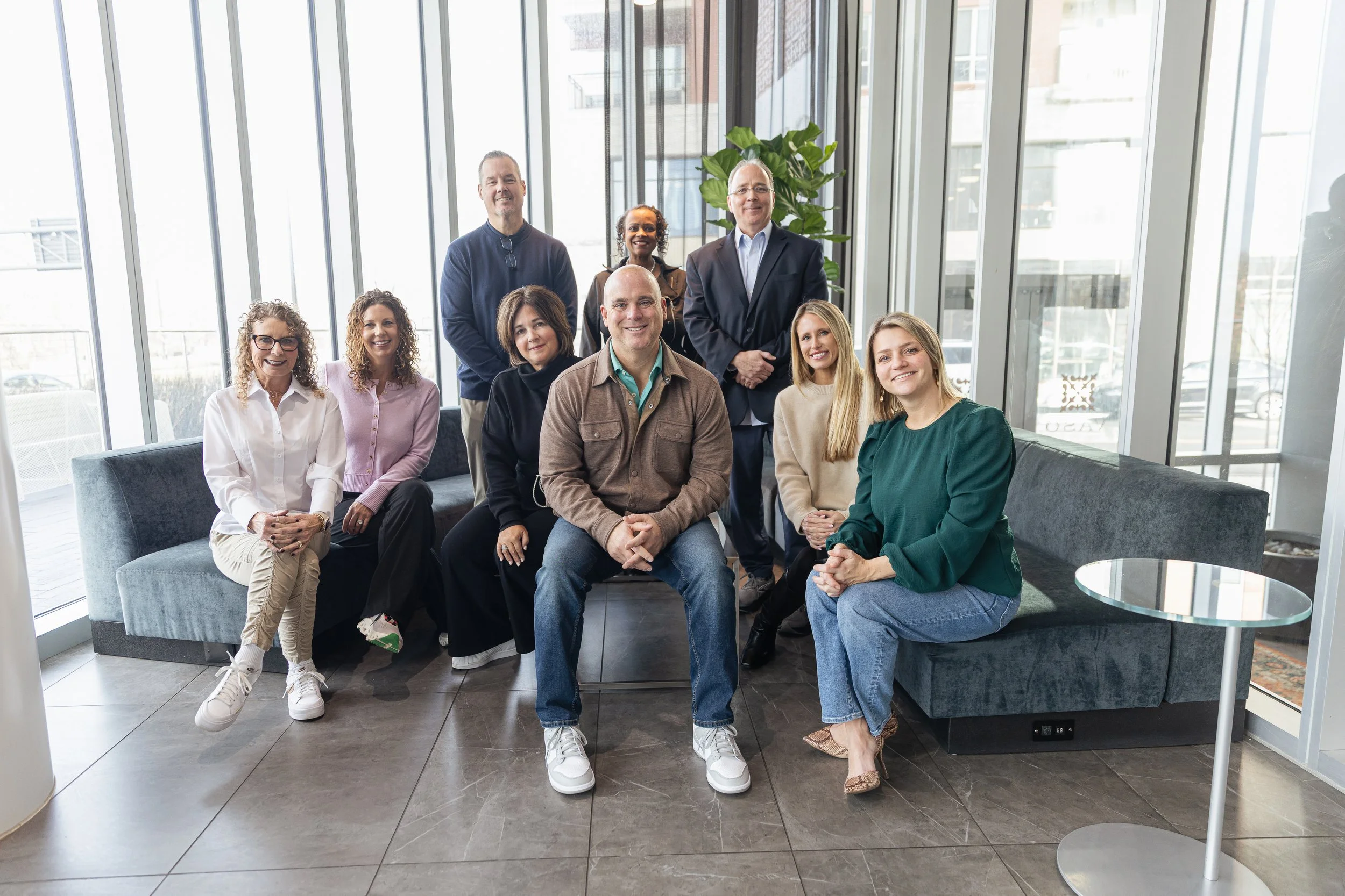 A diverse group of nine people gathered in a modern, glass-enclosed office space, smiling for a group photo.