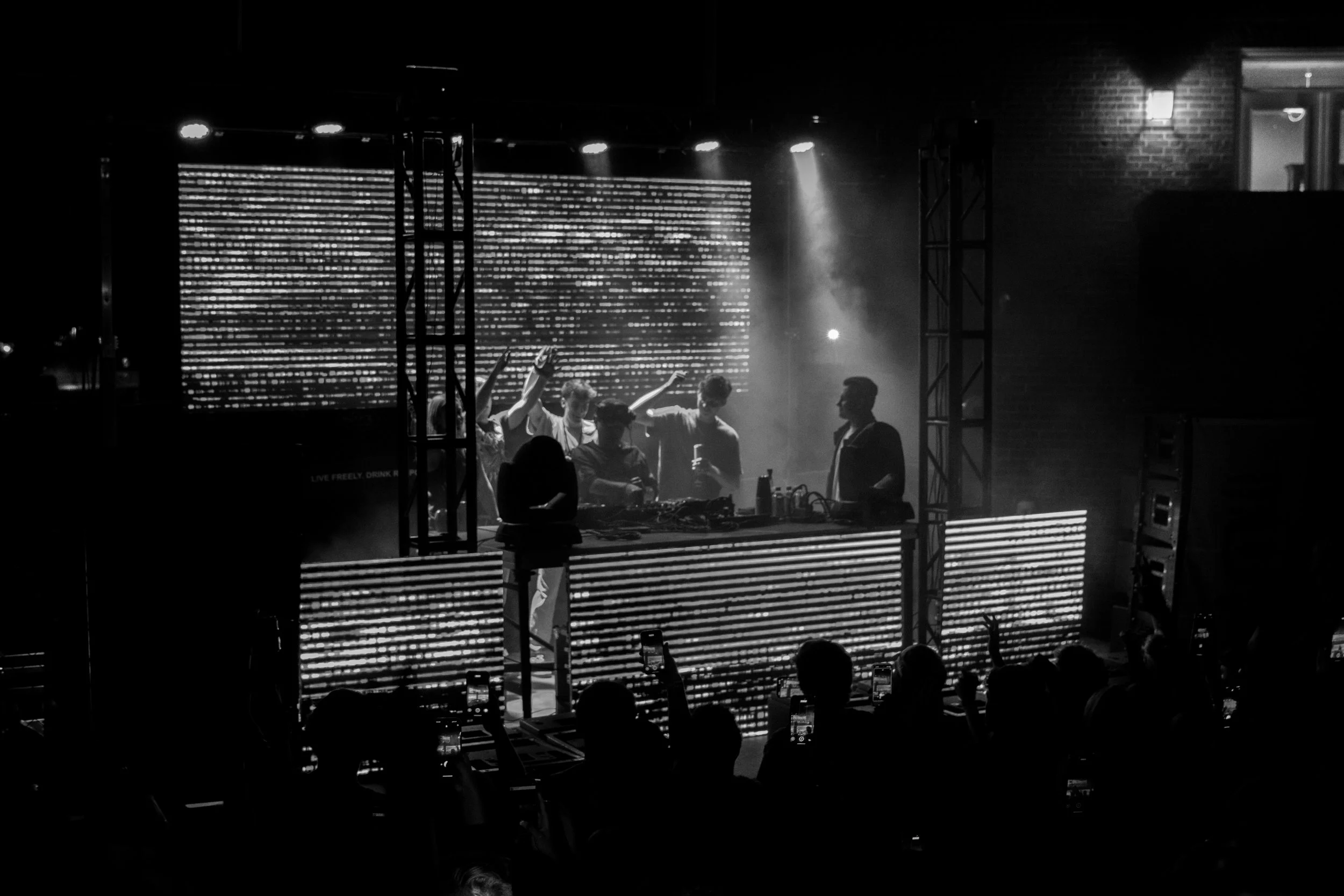 Black and white photo of a DJ performing on stage at a nightclub or concert, with a crowd watching and taking photos. The DJ is behind a large illuminated console with LED screens.