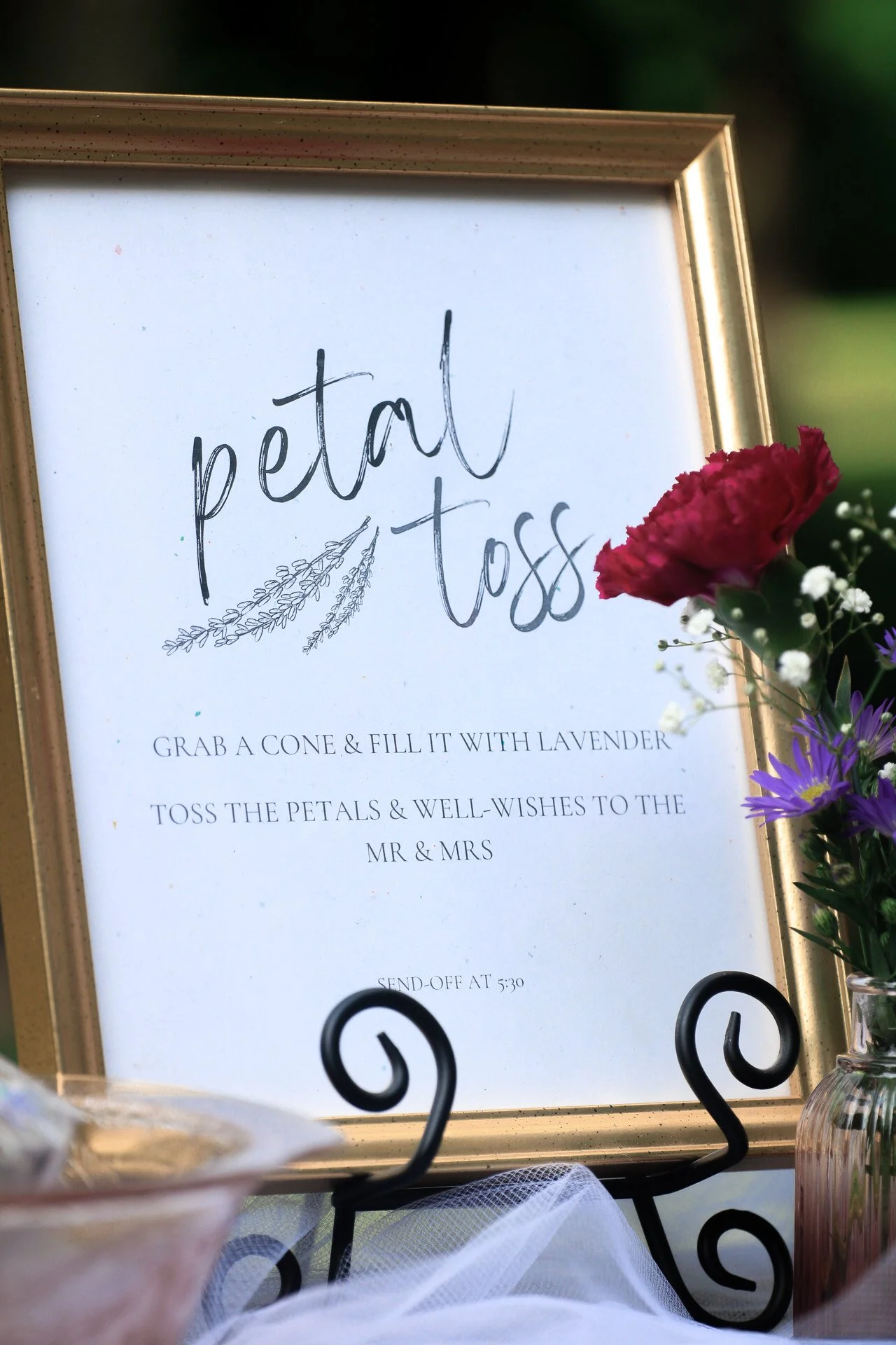 Wedding sign for petal toss with instructions to grab a lavender cone, toss the petals, and well-wishes to the couple, positioned beside flowers in a vase.