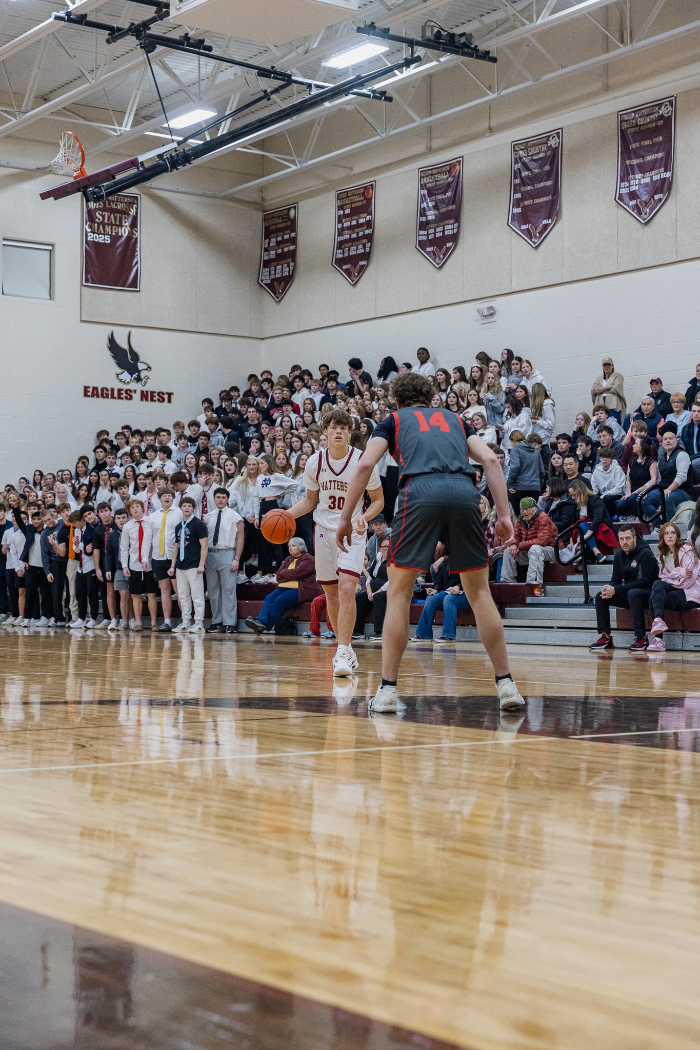 A basketball game is taking place in a school gymnasium filled with students and spectators. Two players are focused on the game, with one in a white uniform dribbling the ball and the other in a dark uniform attempting to block. The gym has banners 