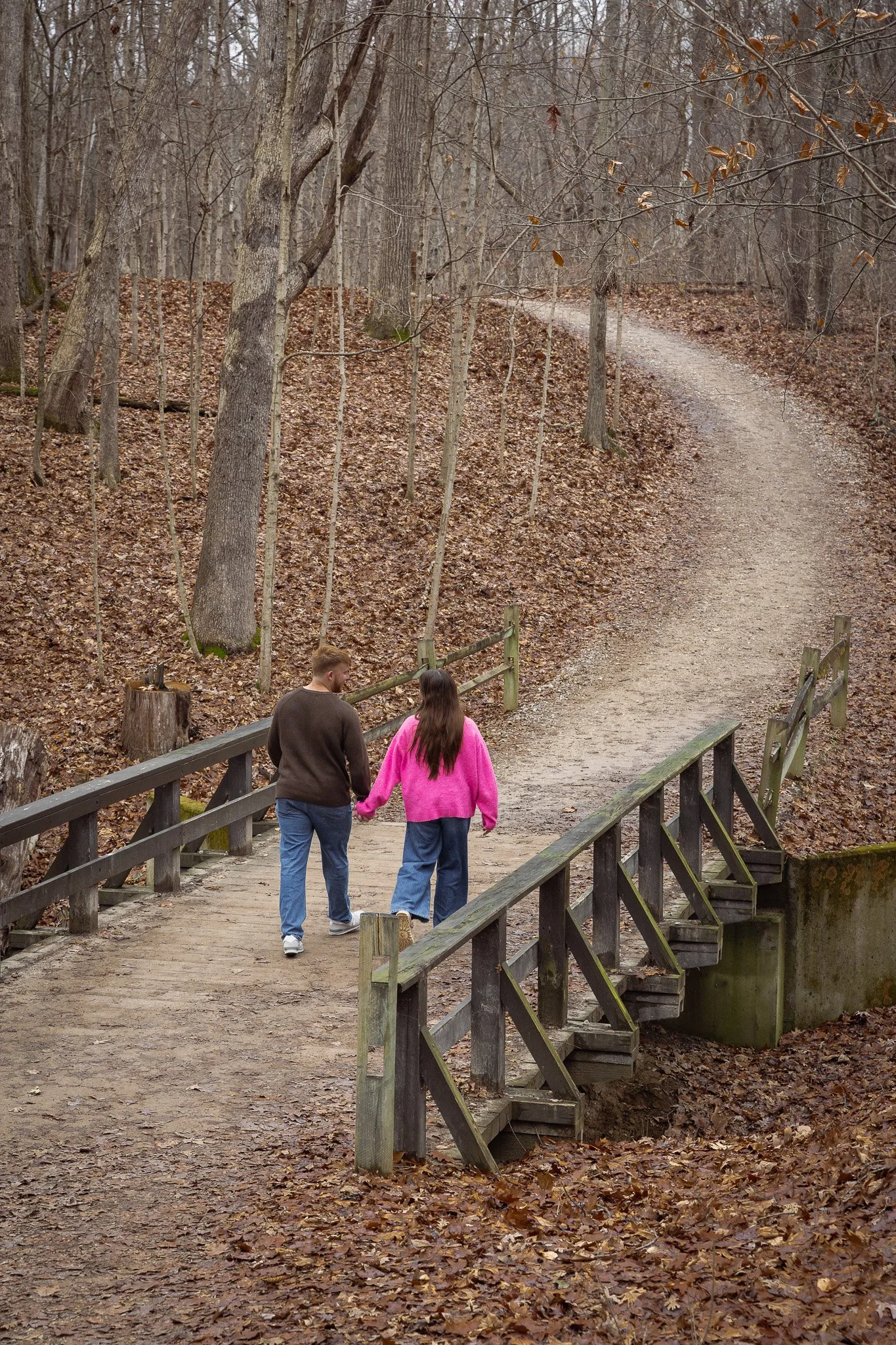 A couple walking hand-in-hand across a small wooden bridge on a dirt trail in a leafless forest during autumn.