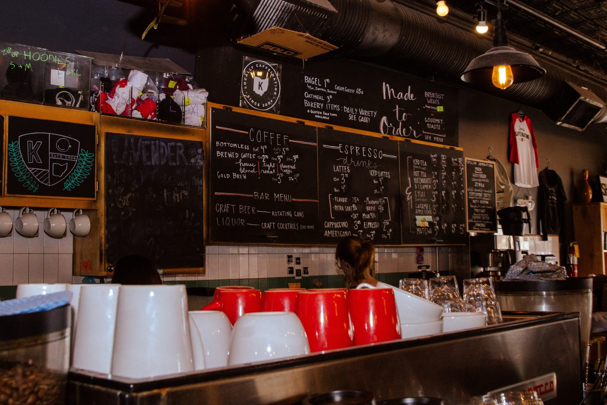 Interior of a coffee shop with black chalkboard menus displaying coffee and drinks, cups stacked on the counter, and warm lighting. Customers are present behind the counter.