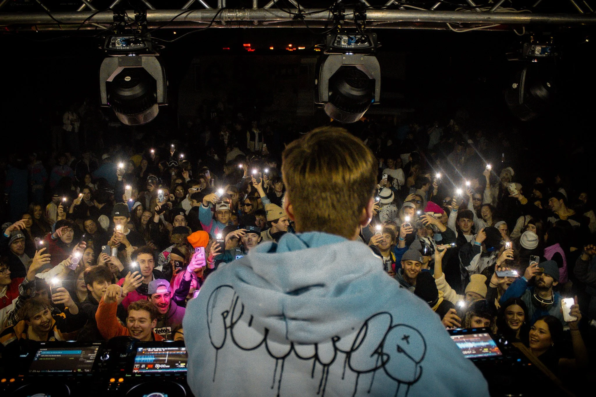 DJ performing at a concert with a large crowd of fans taking photos and videos, many with their phones flashlights on.