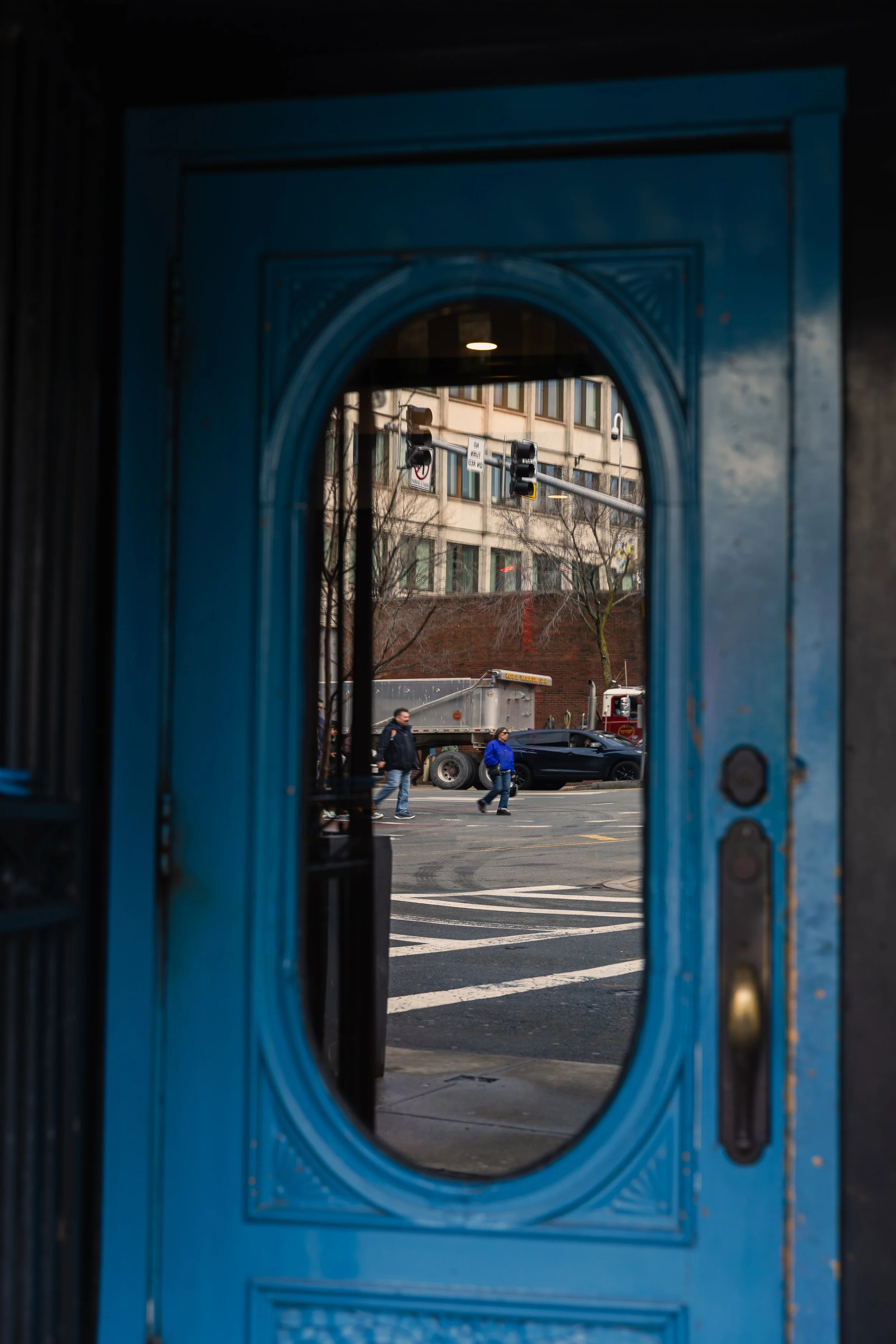 View of a street scene through a blue door's oval glass window, showing pedestrians crossing and vehicles including a black sports car and a delivery truck, with buildings and traffic lights in the background.
