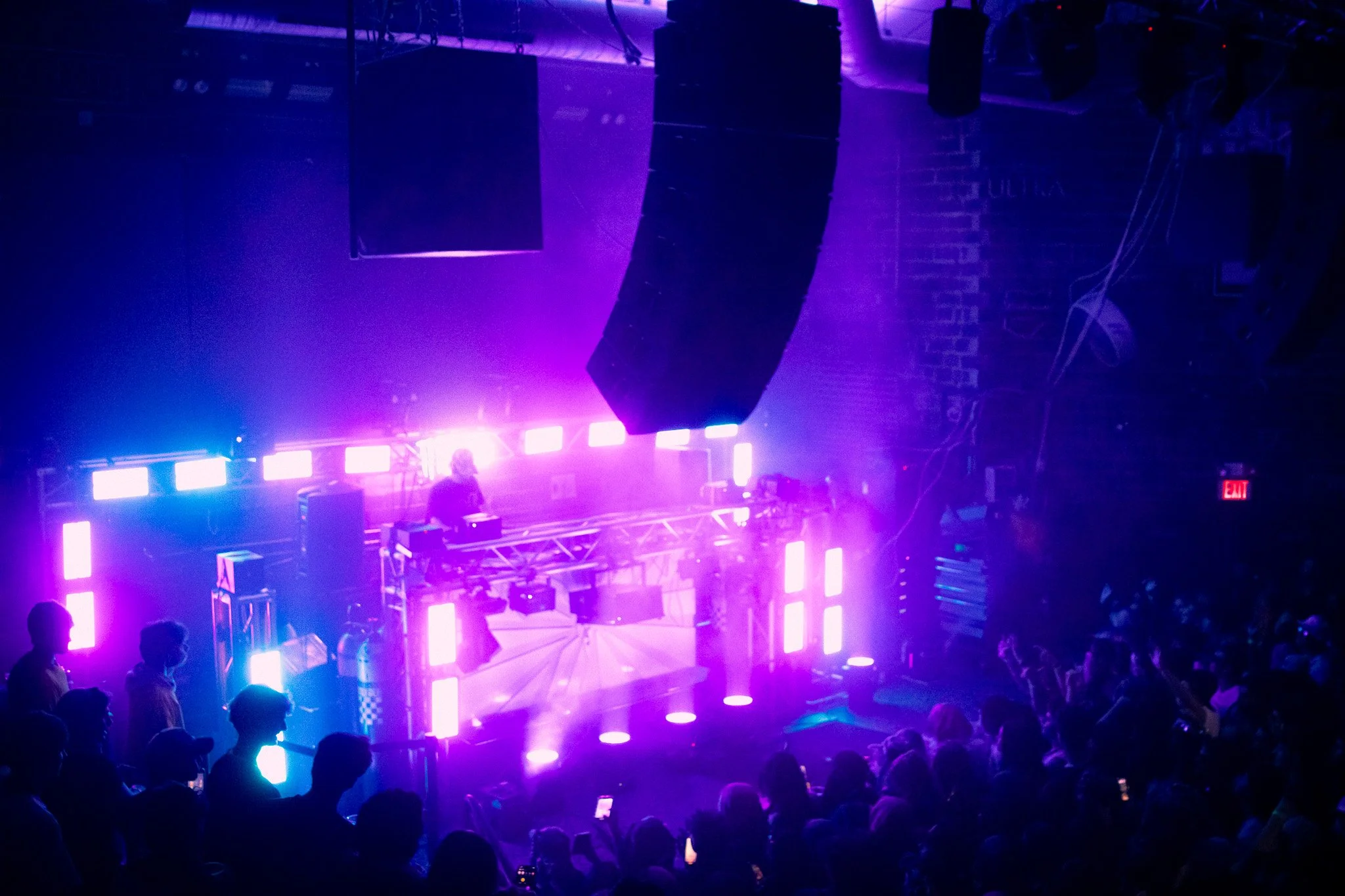 A DJ performs at a nightclub with vibrant purple and blue LED lighting, a crowd watching, and large speakers hanging from the ceiling.