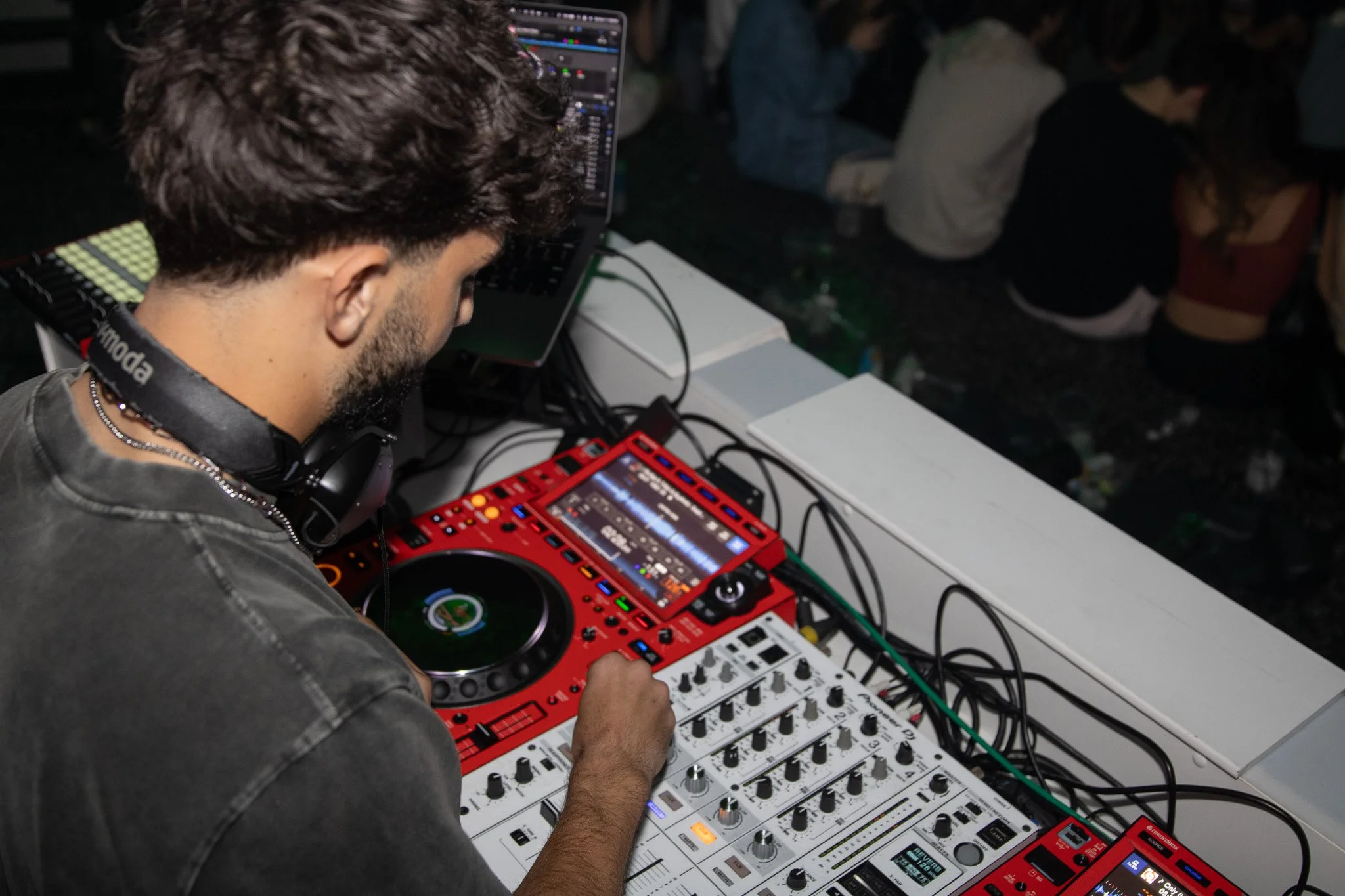 A young male DJ wearing headphones around his neck and a gray shirt, operating a DJ controller with a red and white color scheme at a party or club. In the background, audience members are seated on the floor, some facing away from the camera.