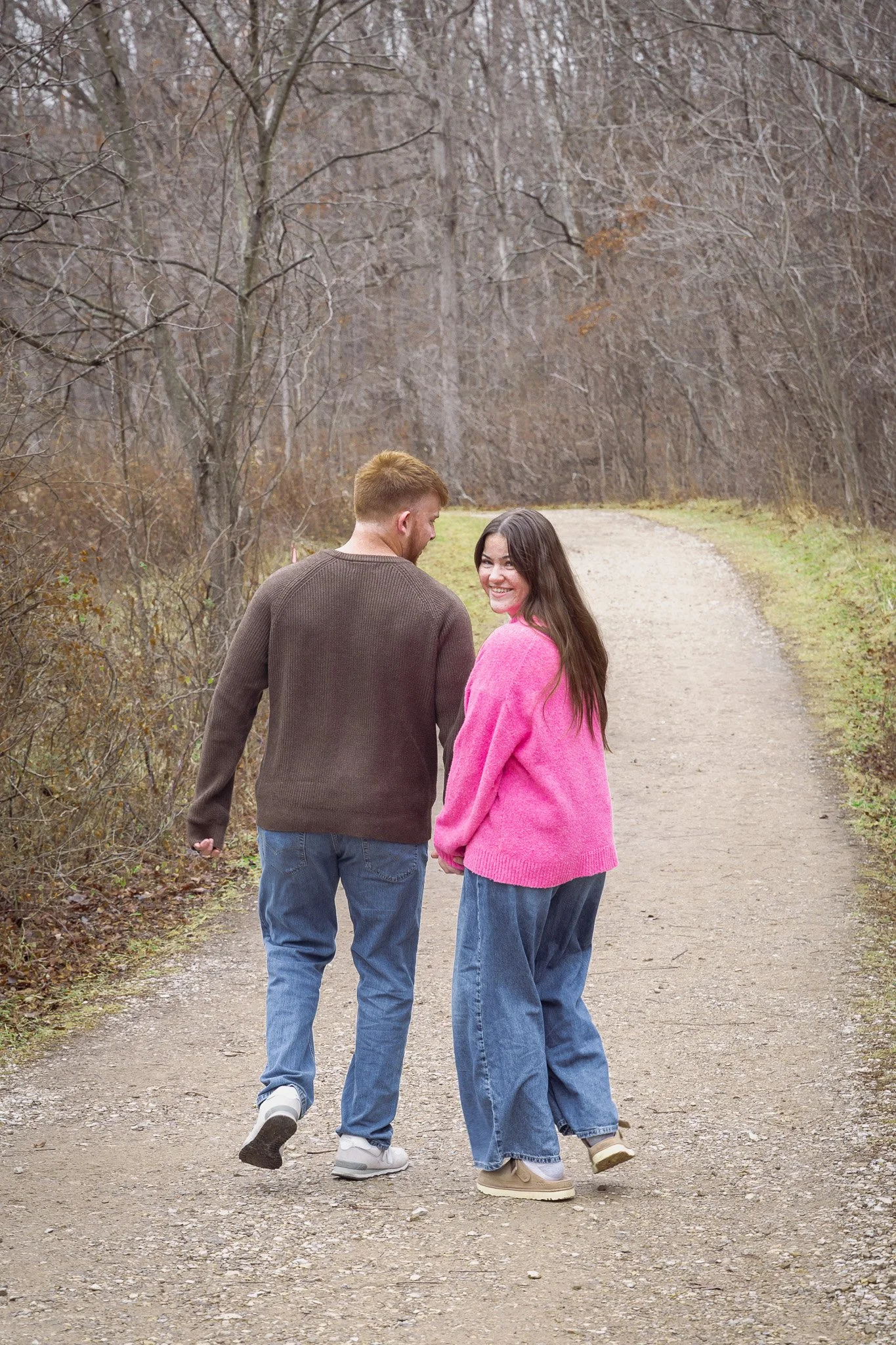 A young couple walking hand in hand on a dirt trail in a wooded area during fall or winter, with the woman smiling and looking back at the camera.