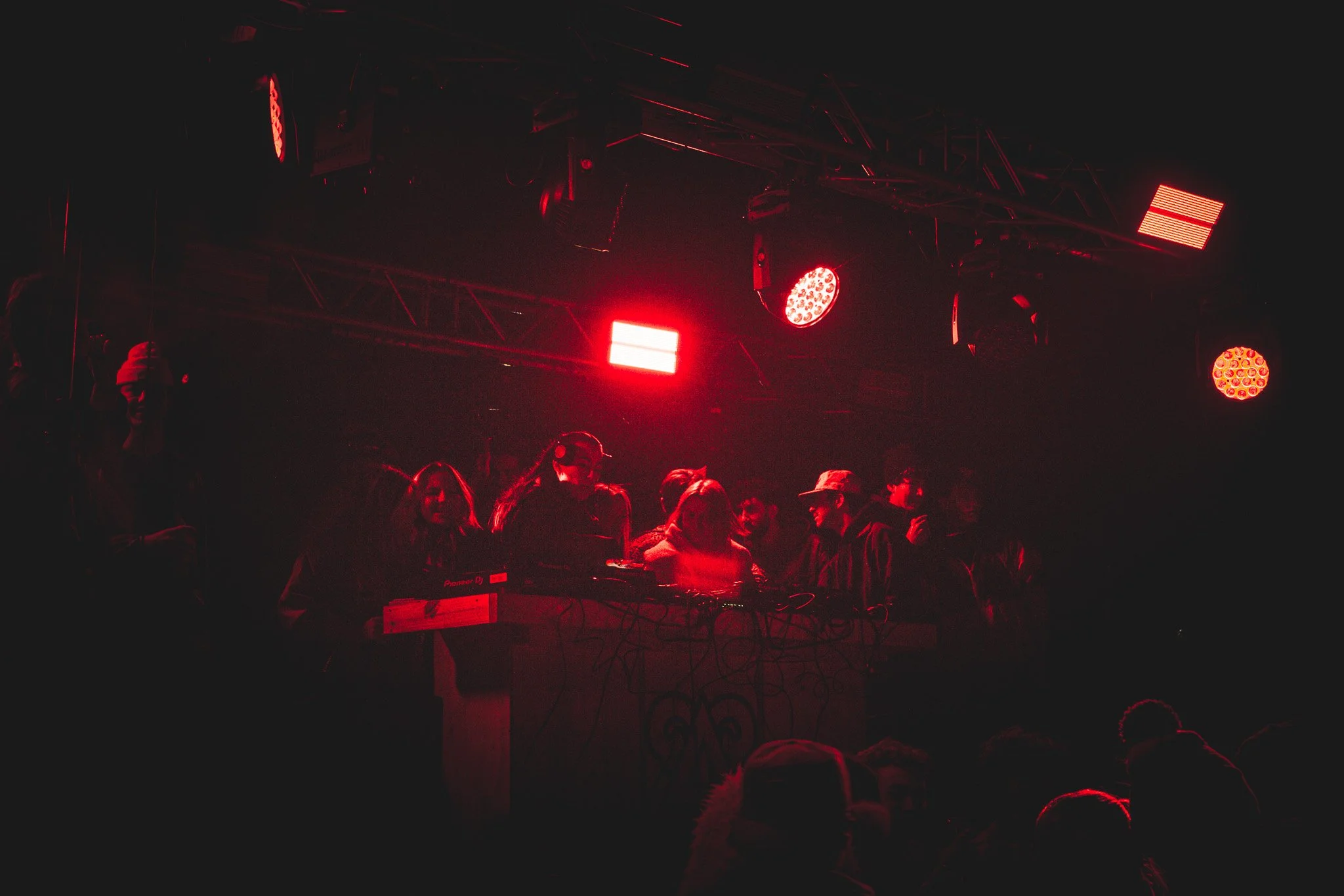 Dimly lit scene of a DJ performing on stage with red and orange stage lights, with people dancing and enjoying the music in front of the DJ booth.