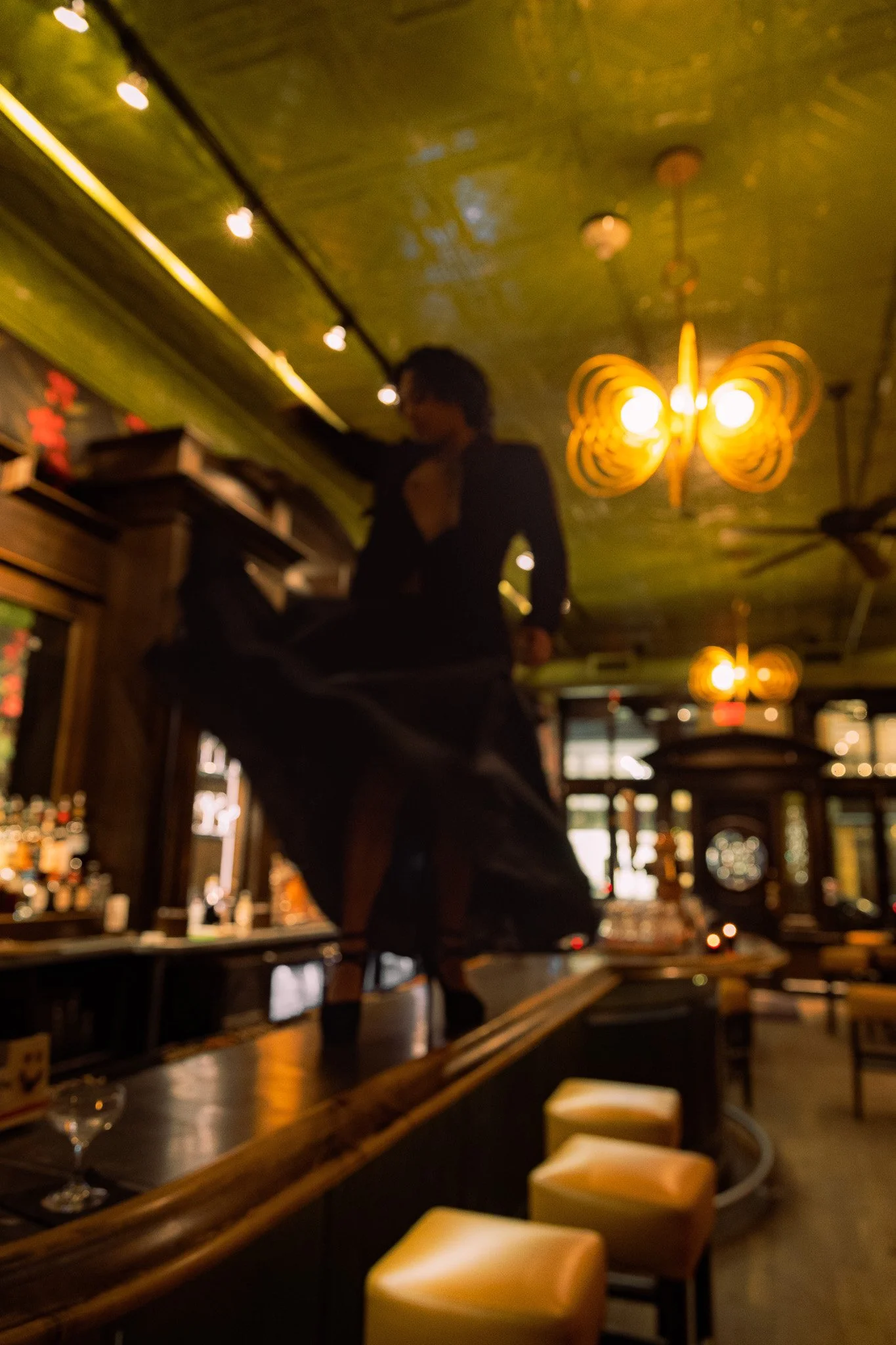 A woman in a black dress standing on a bar counter inside a dimly lit restaurant with warm lighting and decorative ceiling fixtures.