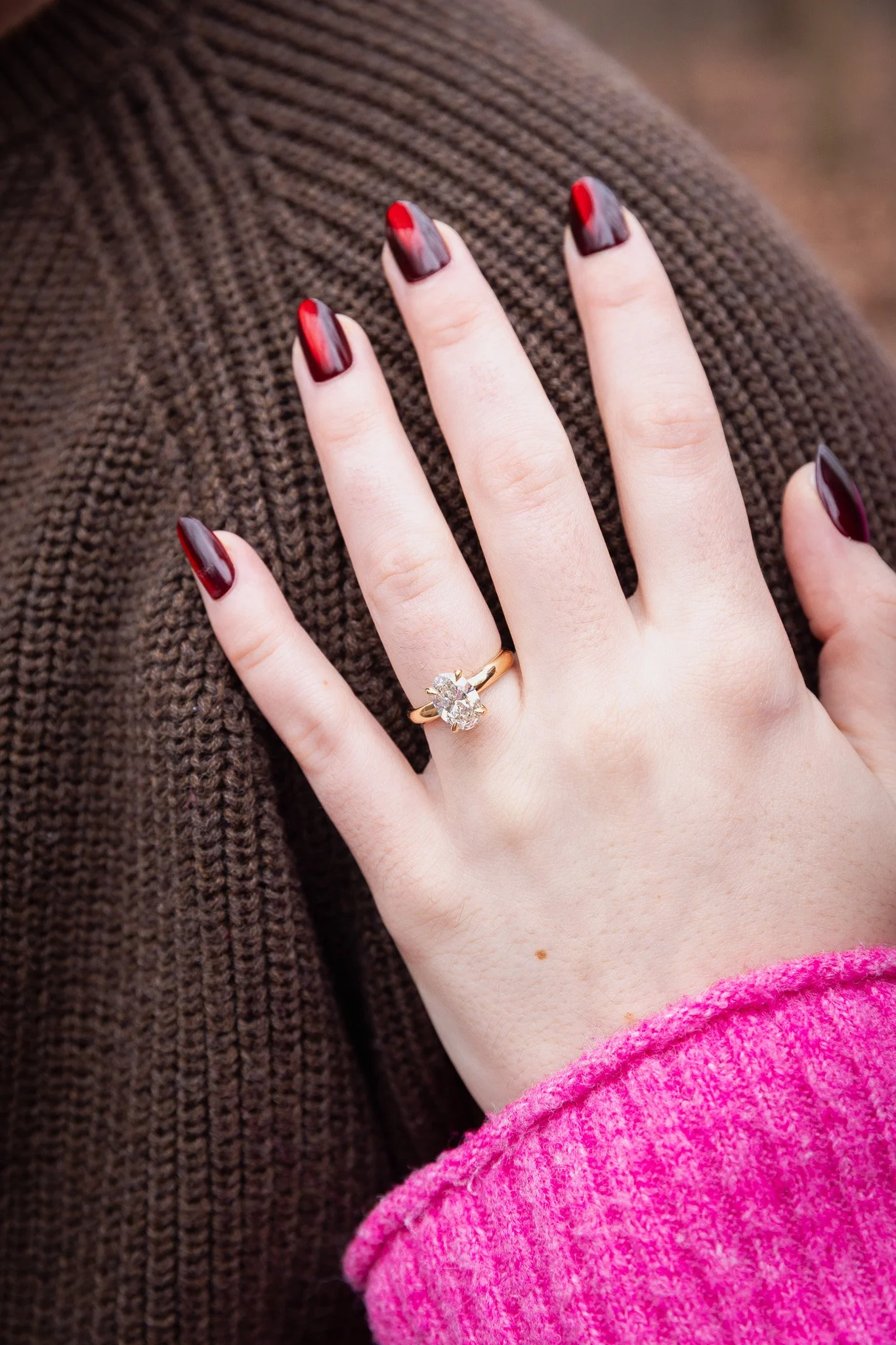 A woman's hand with dark red and gray nail polish, wearing a gold ring with a large gemstone, resting on a brown knitted fabric.
