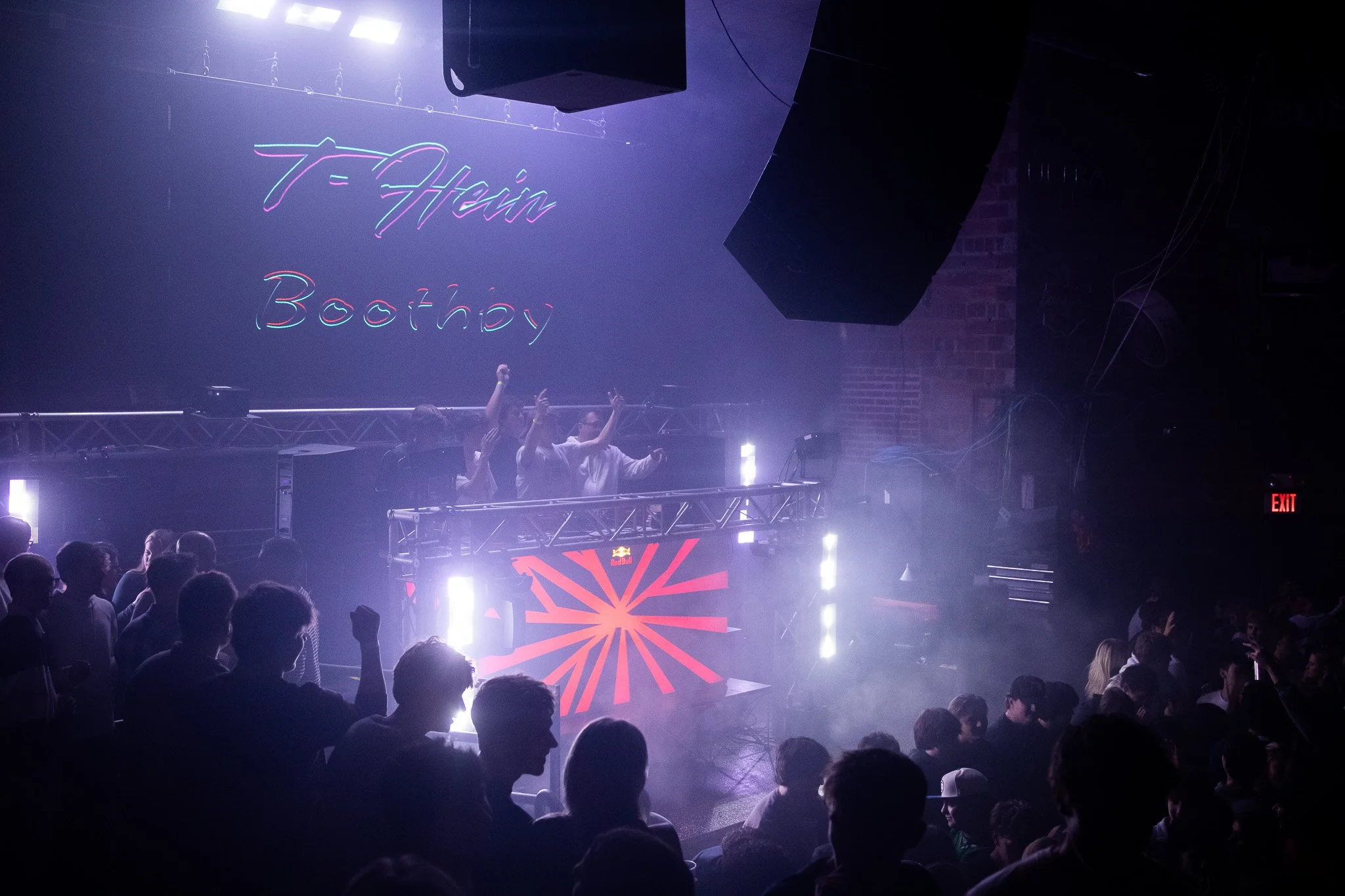 Crowd dancing at a nightclub with a DJ booth, neon sign reading 'T-Hain Boothby', and colorful lights.