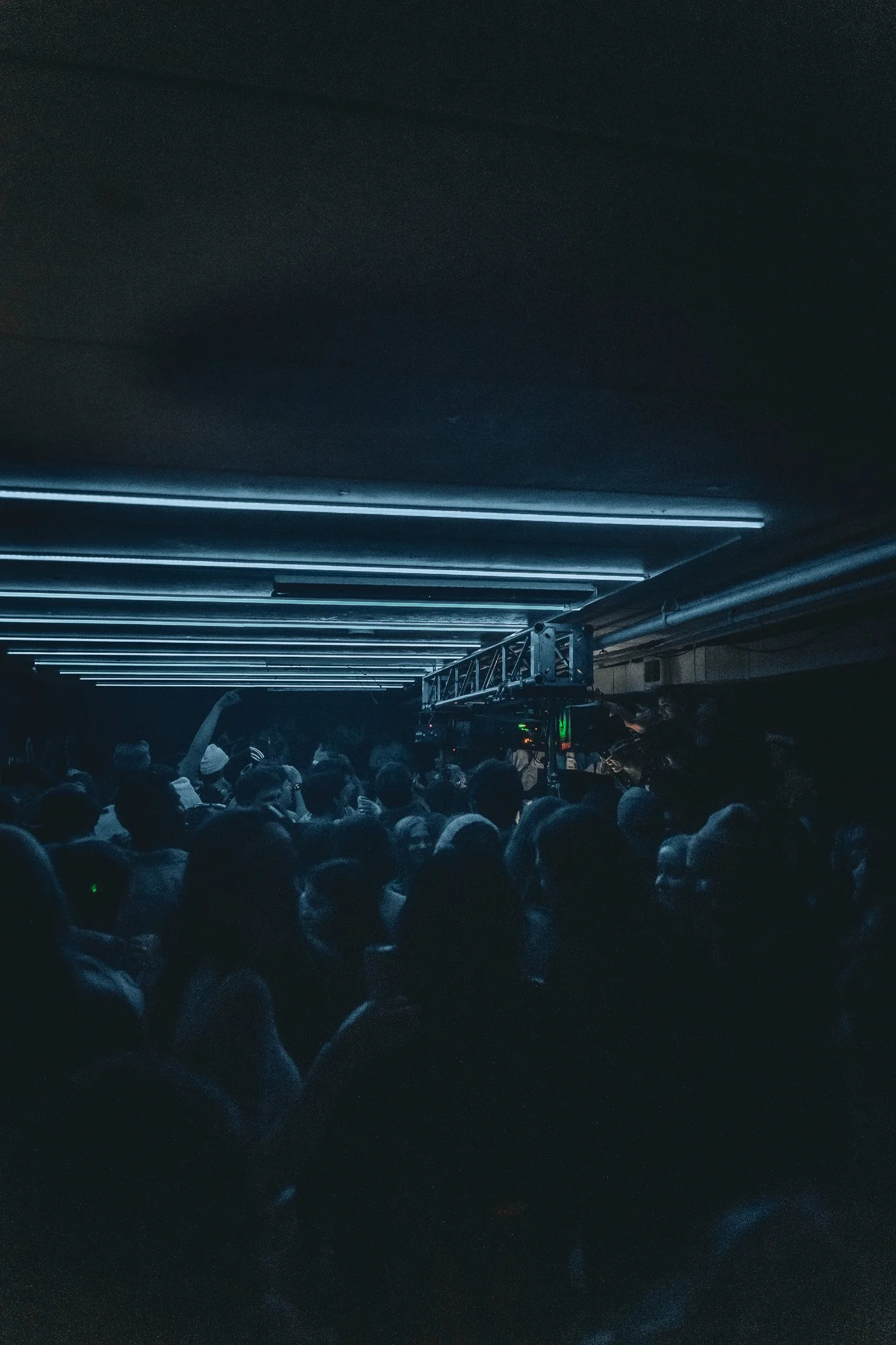 Crowd of people at a dark indoor concert or event with blue lighting and a metallic structure overhead.