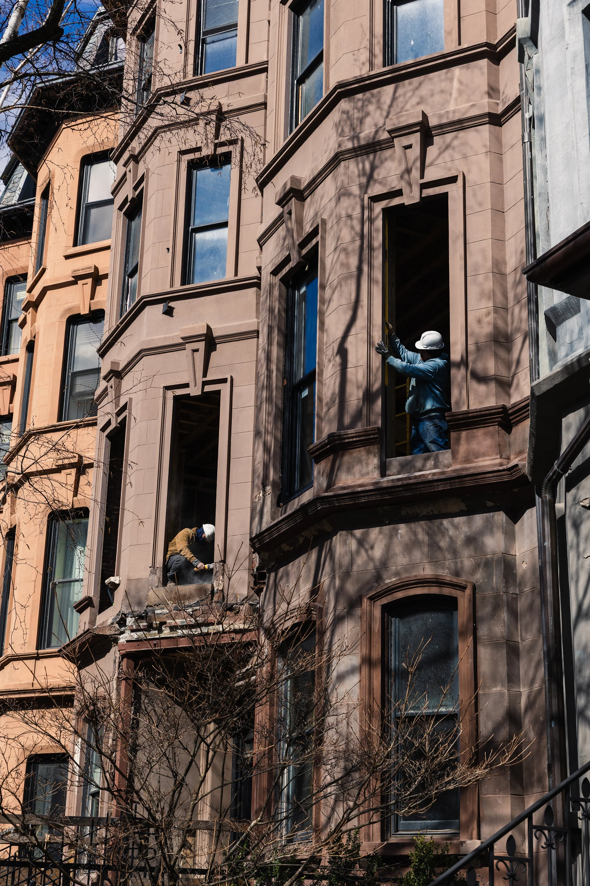 Construction workers wearing helmets working on a brownstone building, with one worker on a ladder and another crouching inside a window opening, surrounded by leafless trees and other similar buildings.