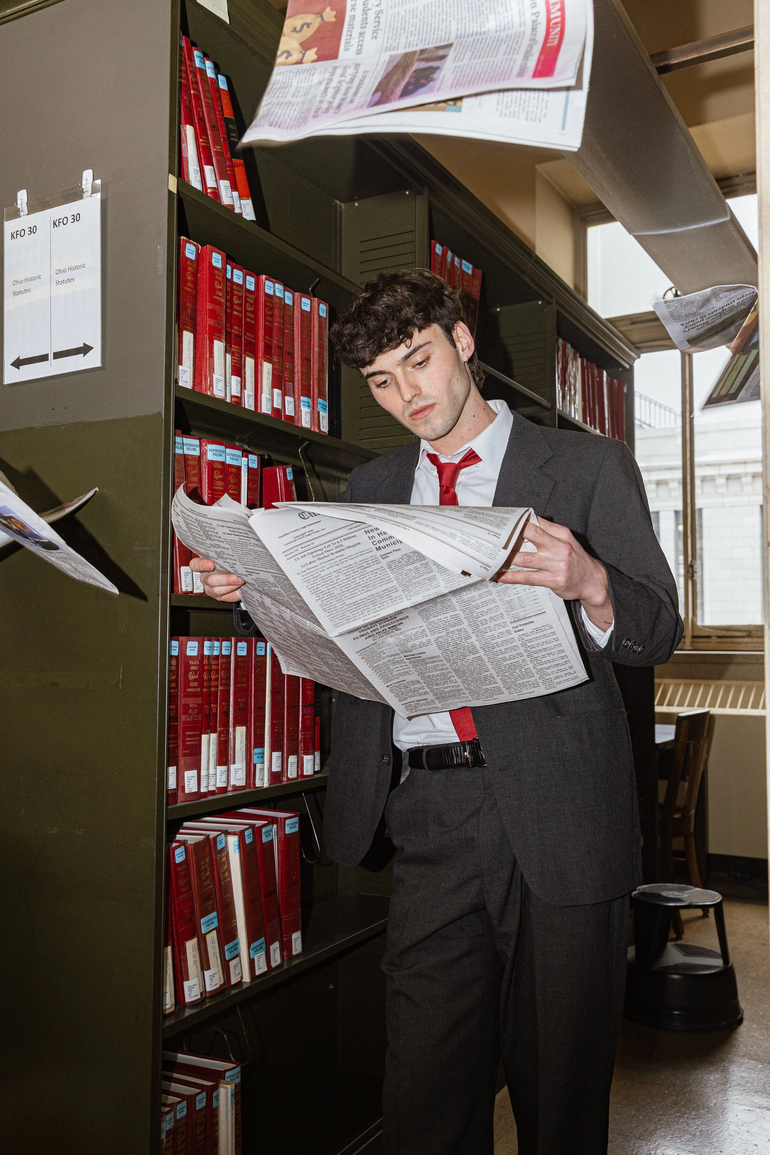 A young man in a dark suit and red tie reading a newspaper in a library filled with red books on shelves.