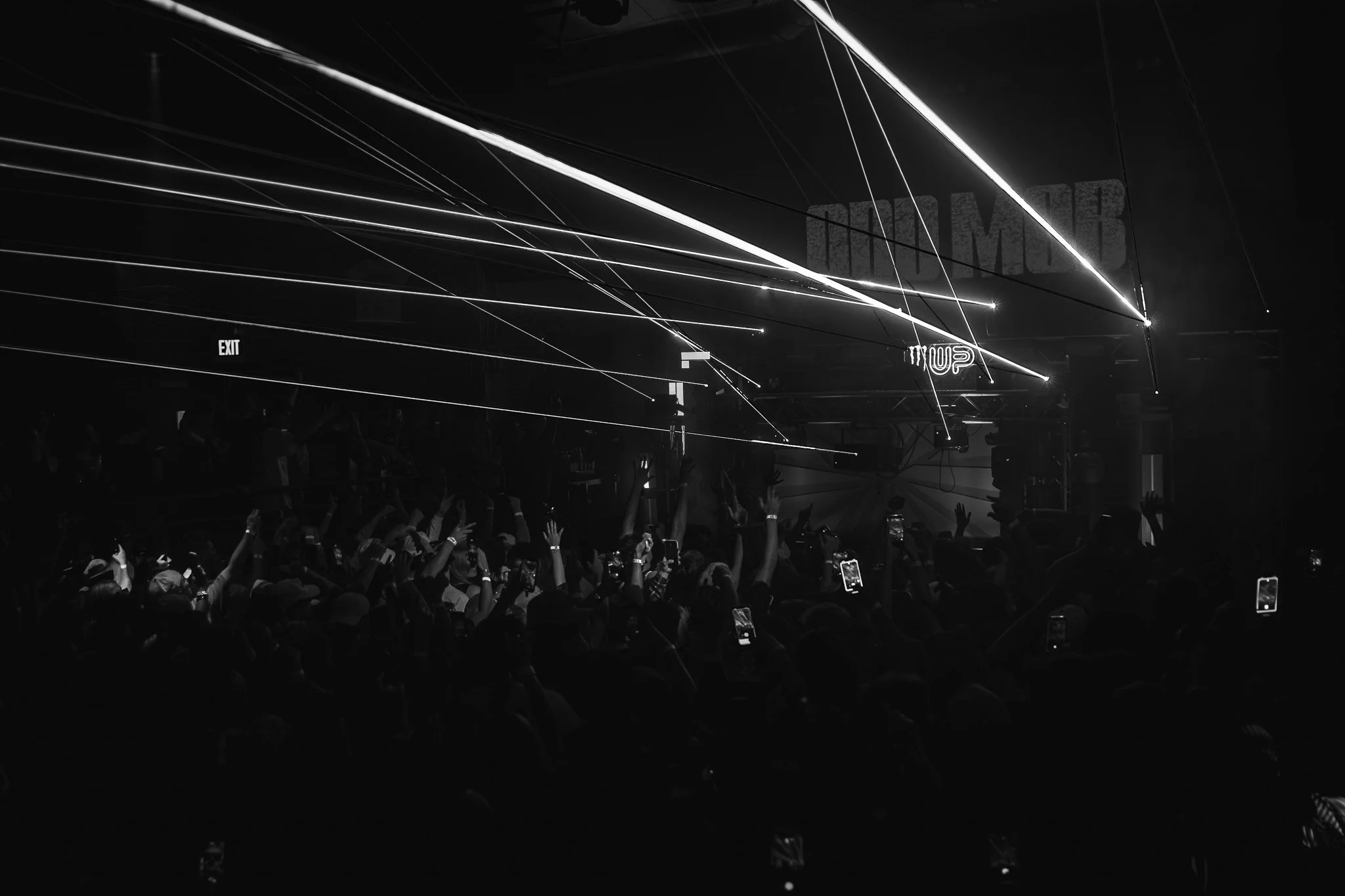 Crowd of people at concert or event with their hands raised, illuminated by sparse lighting and laser effects, dark setting with visible exit sign.