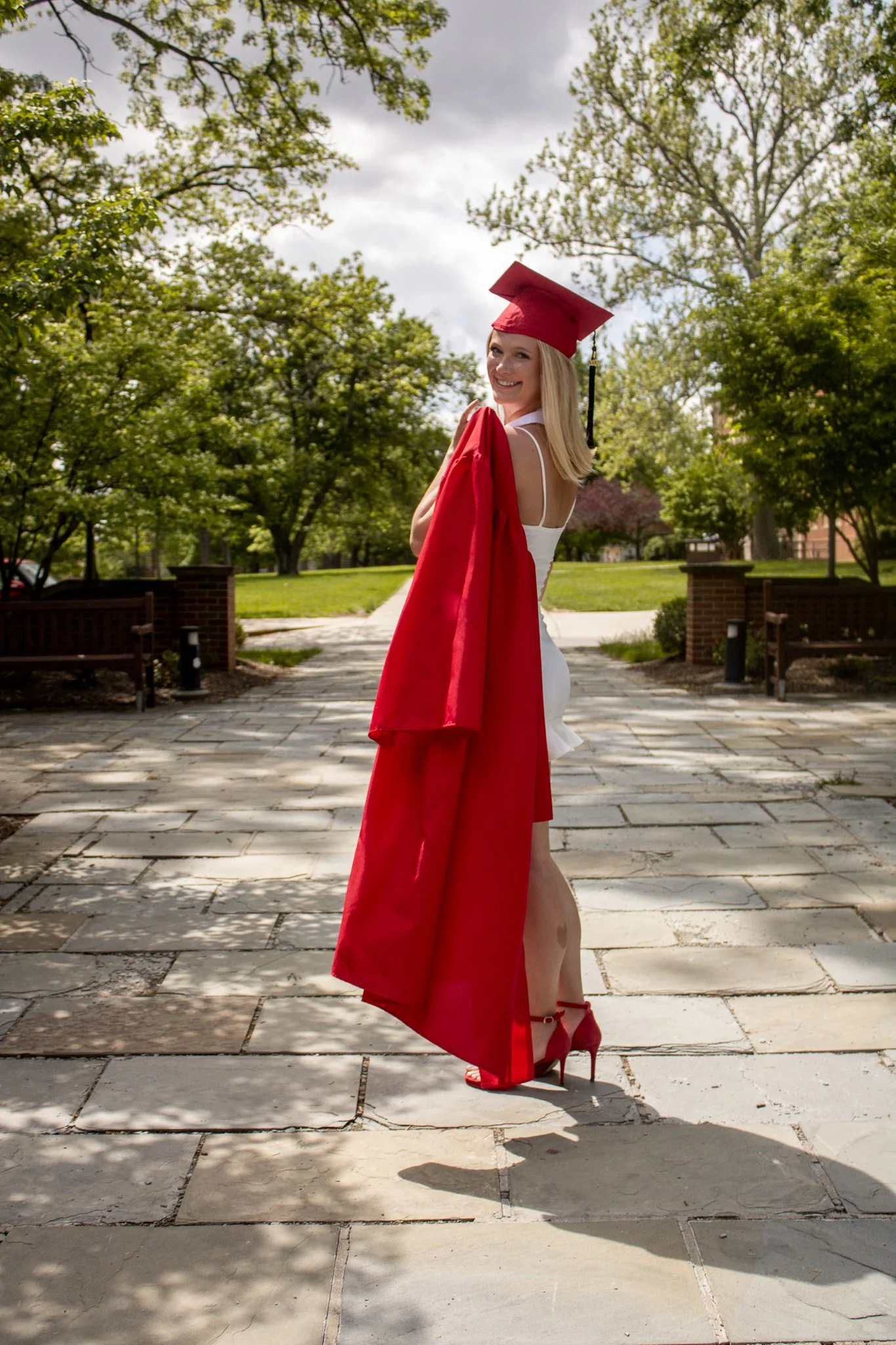 Young woman in a white dress with red high heels, red graduation cap, and gown draped over her shoulder, standing outdoors on a stone pathway with green trees and a cloudy sky in the background, smiling at the camera.