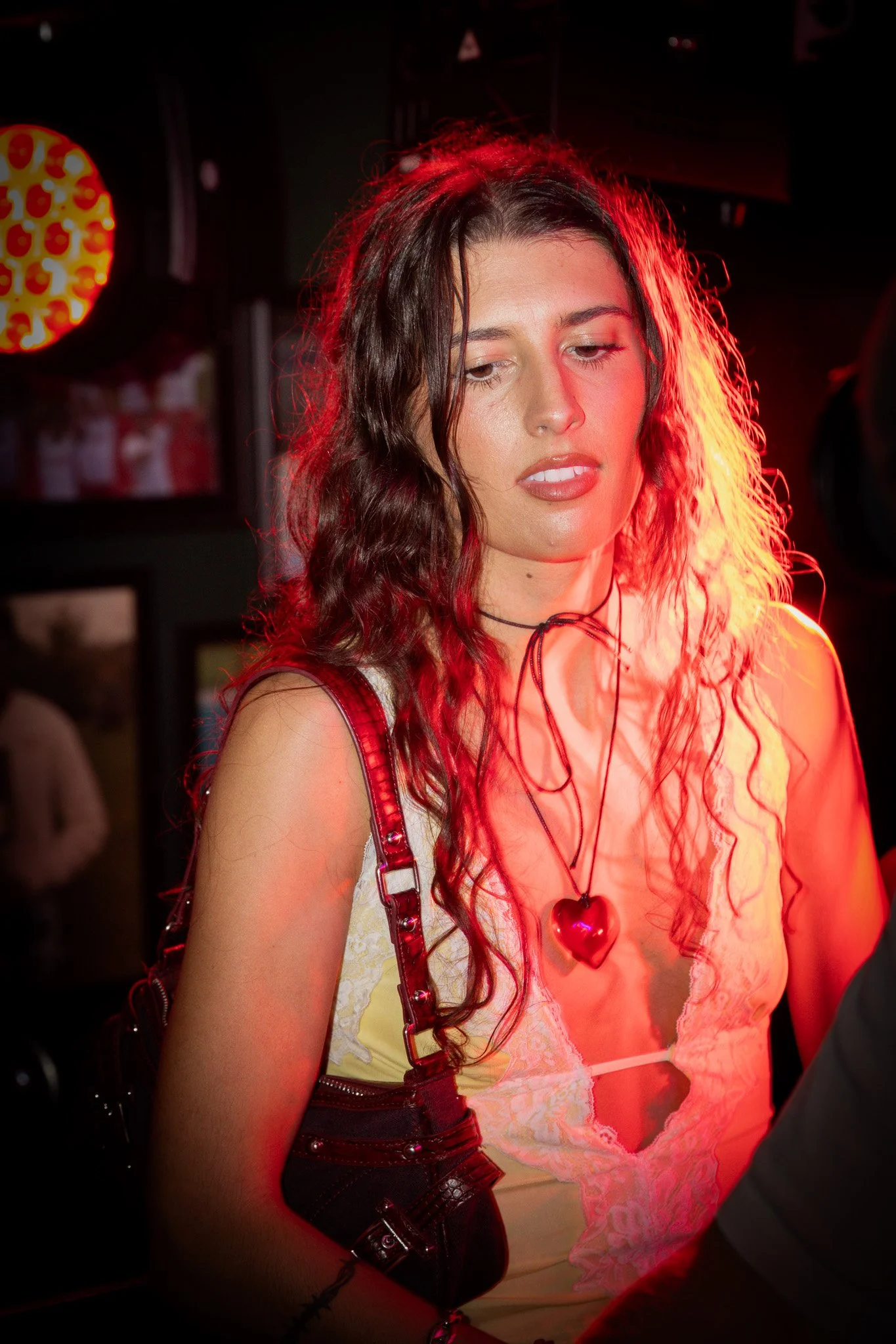 A woman with long, curly brown hair wearing a yellow dress with lace detail and a heart-shaped necklace, illuminated by red lighting in a dimly lit venue.