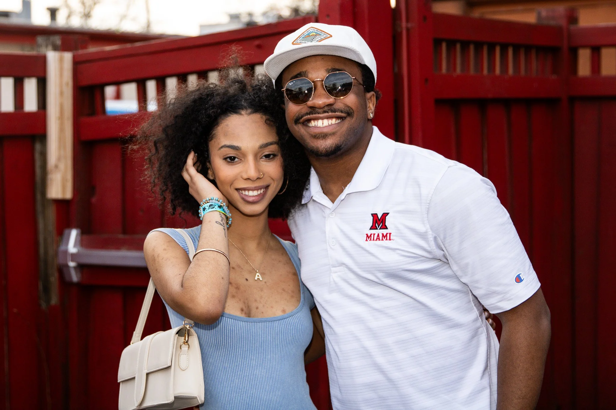 A smiling couple poses arm-in-arm outdoors in front of a red wooden fence. The woman has curly black hair, wears a sleeveless blue top, jewelry, and carries a beige purse. The man wears sunglasses, a white cap, and a white polo shirt with a Miami log