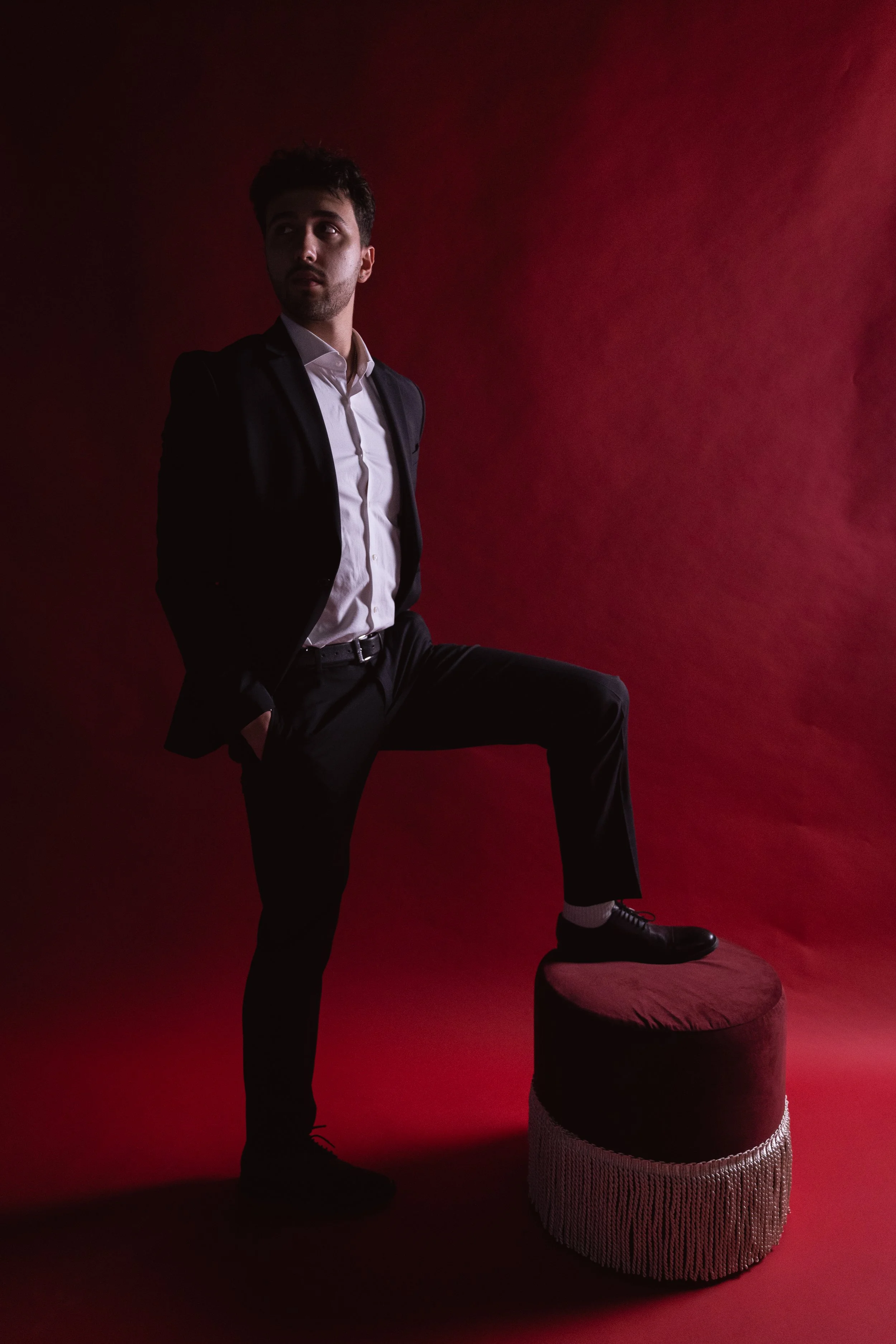 A man in a black suit with a white shirt standing with one foot on a velvet ottoman against a red background.
