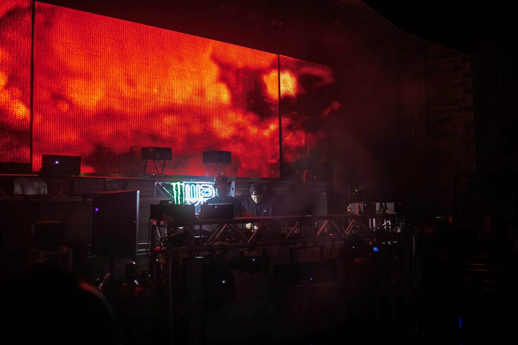 A DJ performing on stage with large LED screens displaying fiery red and orange visuals behind him, in a dark indoor venue.