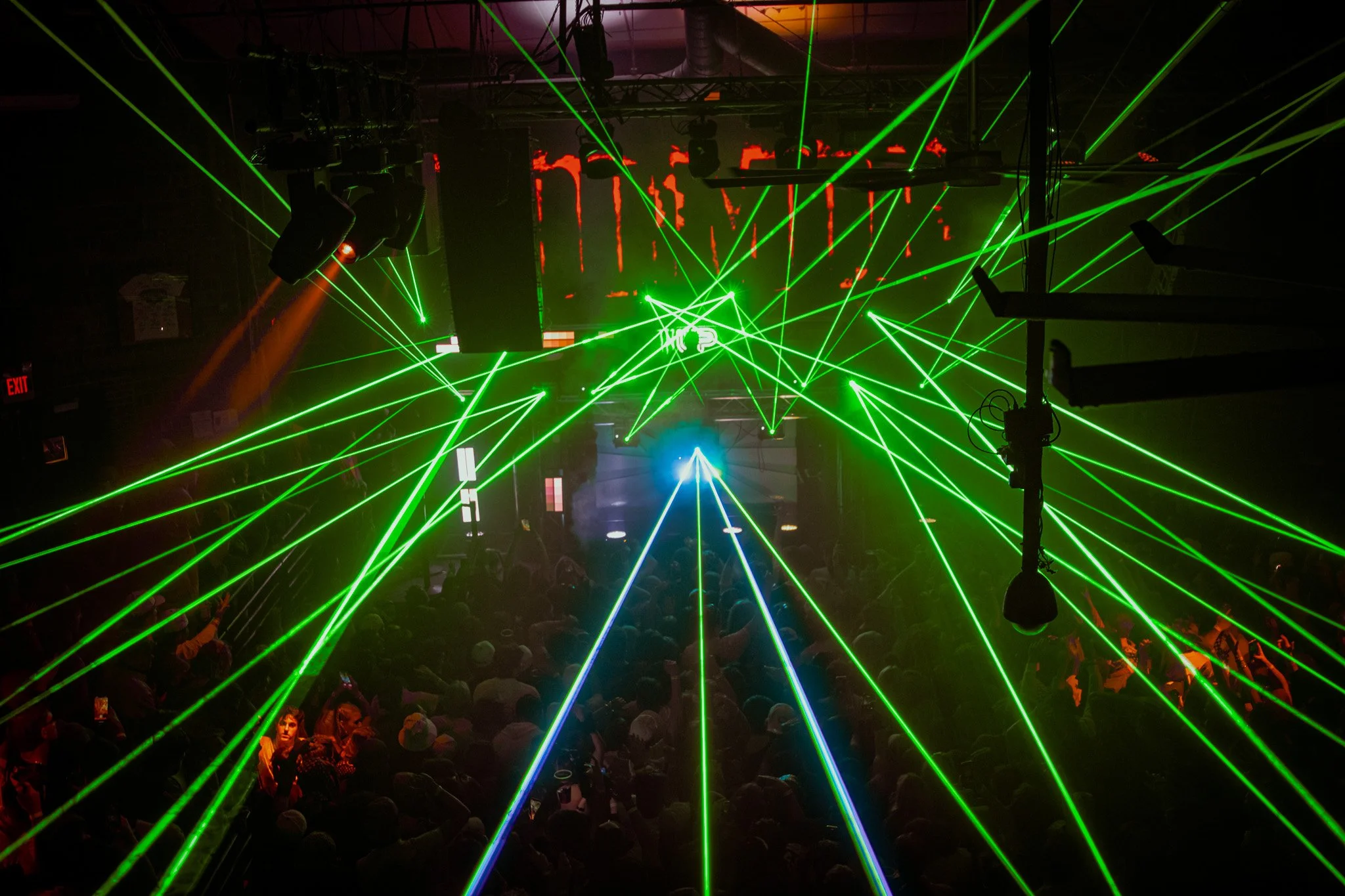 Laser lights in green and blue crossing over a crowd at a concert or nightclub, with a stage and a screen displaying red graphics in the background.
