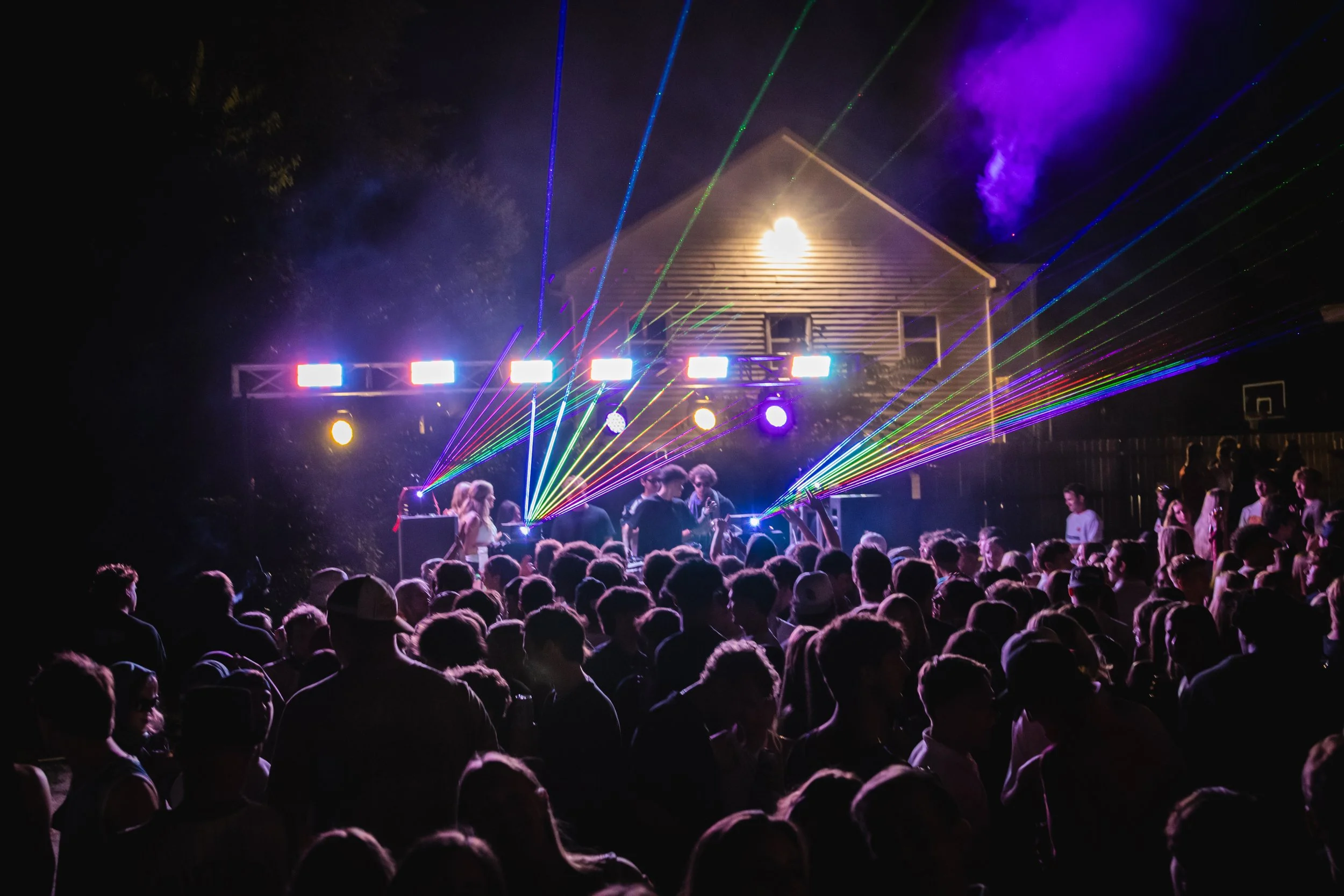Crowd of people gathered outdoors at night for a concert with colorful laser lights and a stage.
