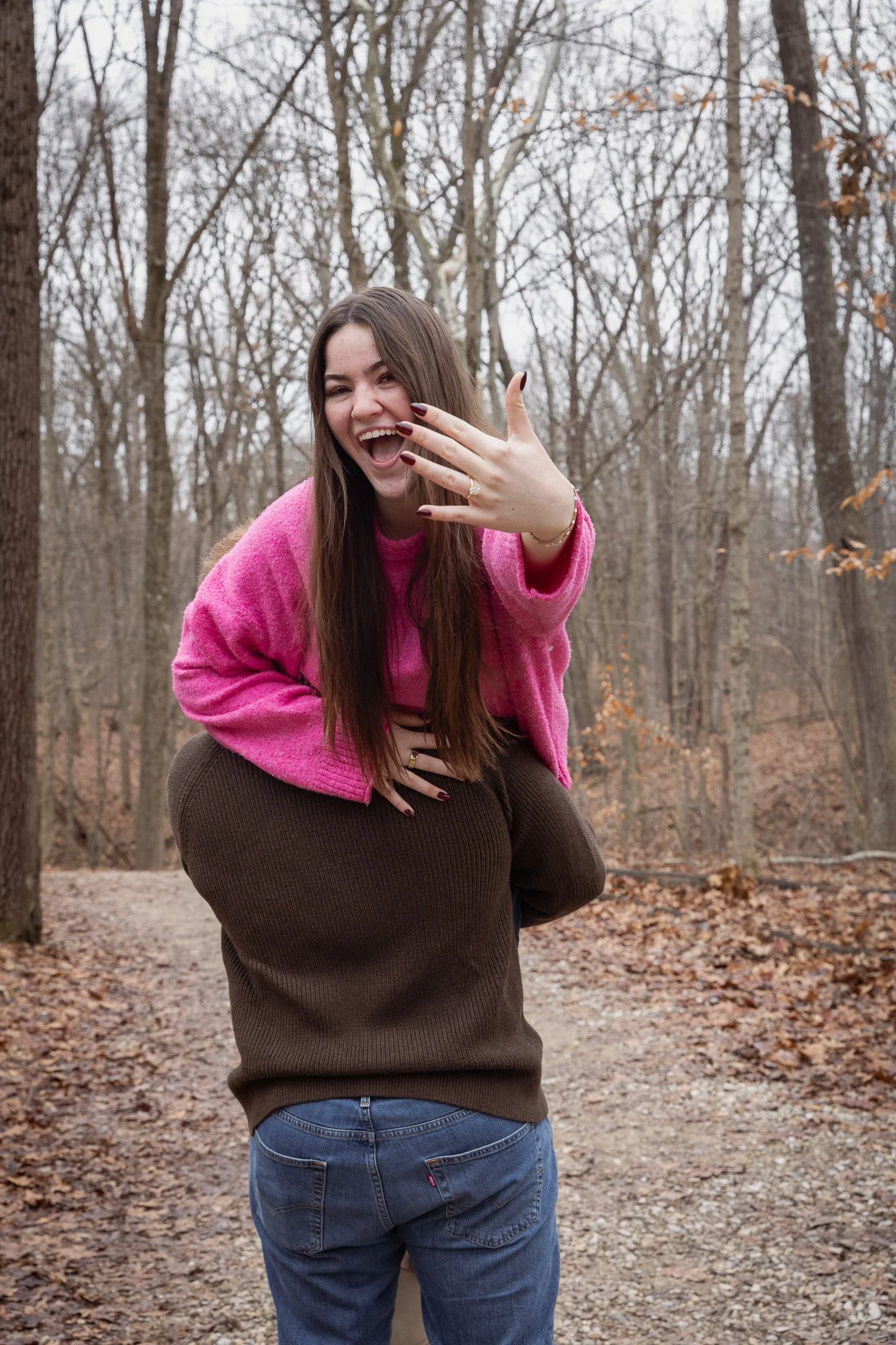 A woman with long brown hair, wearing a pink fuzzy sweater and blue jeans, is smiling and showing her hand with rings, while crouching on a trail in a wooded area during autumn.