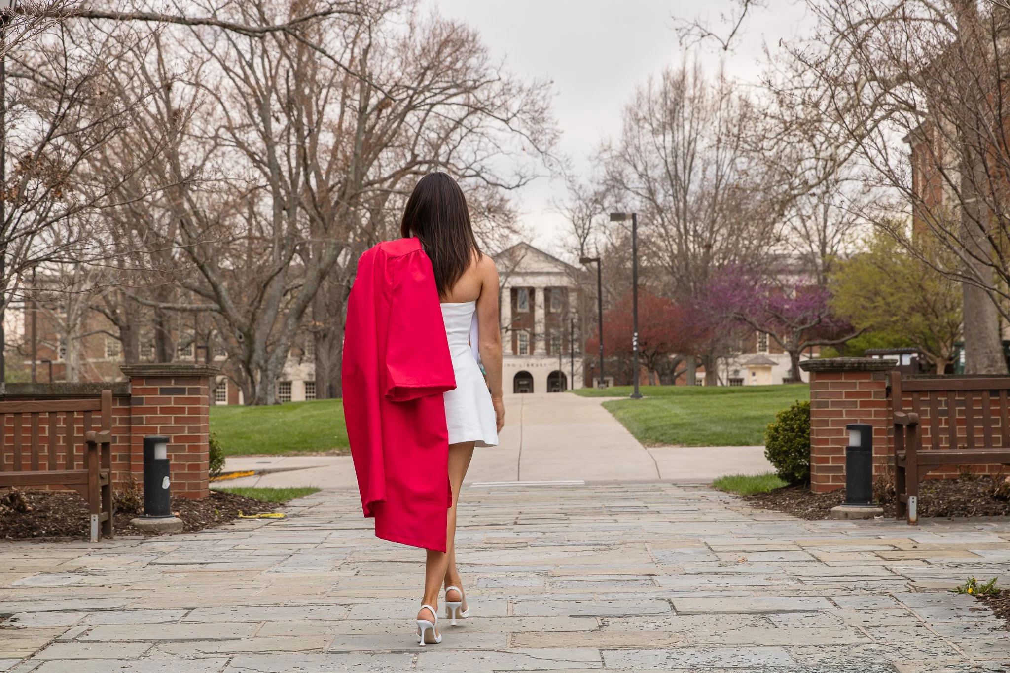 A woman in a white dress and high heels walking away on a stone pathway, carrying a bright pink garment over her shoulder, with a university building and trees in the background.