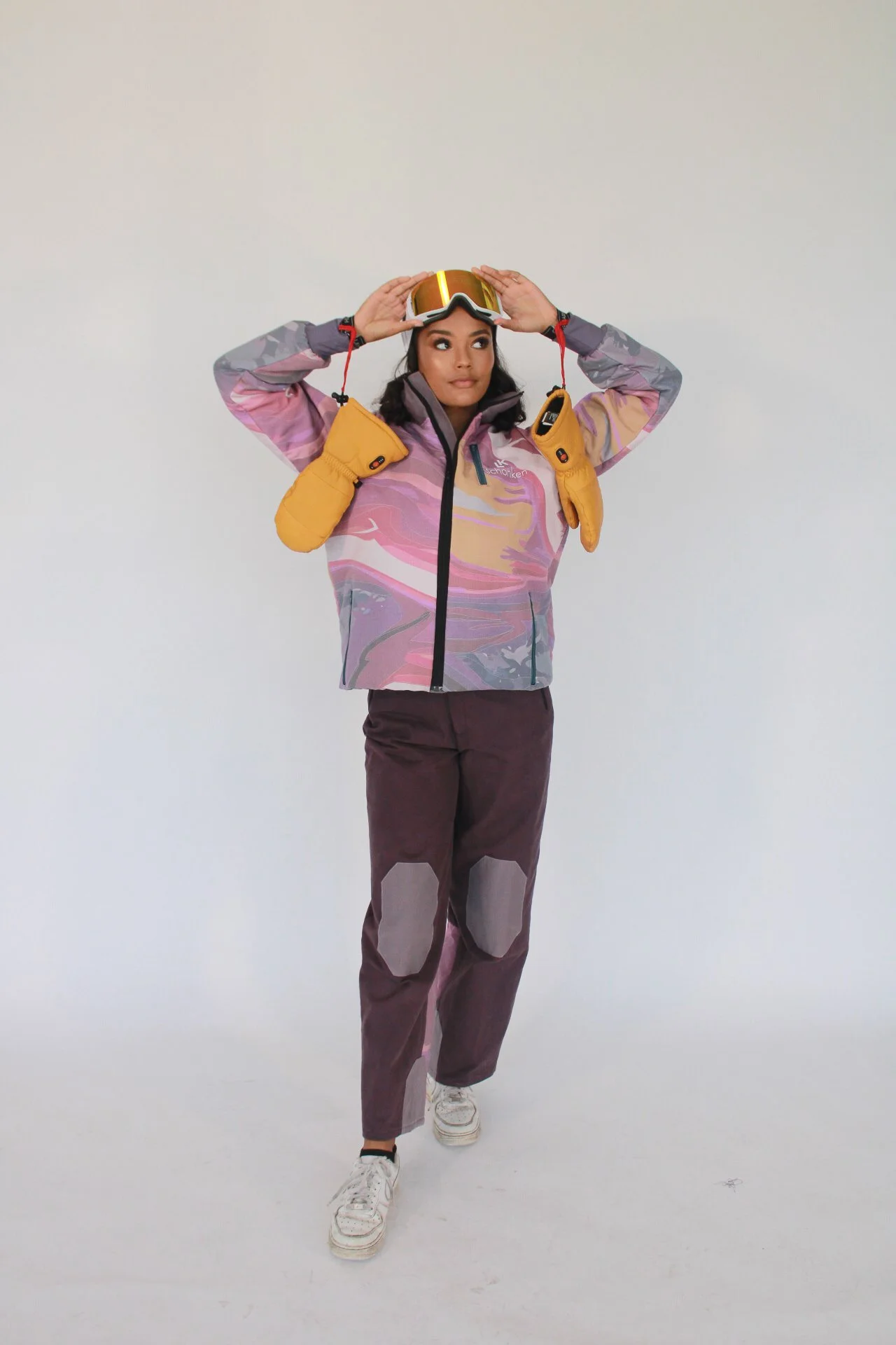 Woman putting on ski goggles, dressed in skiing gear including a colorful jacket, pants with knee patches, yellow mittens, and white sneakers, against a plain white background.