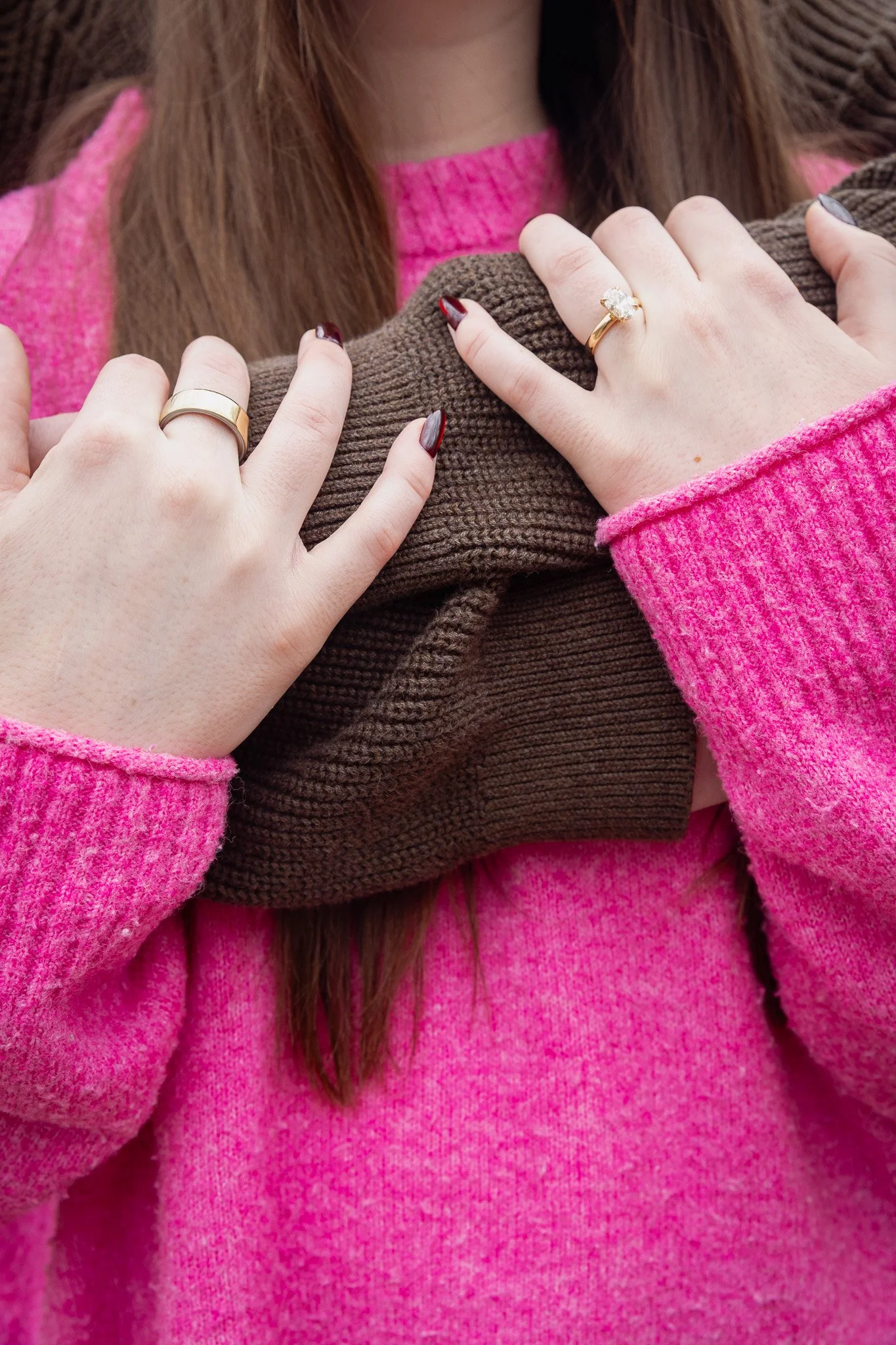 Close-up of two women wearing rings; one with a gold band and the other with a large engagement ring, embracing each other. Both wearing pink and brown sweaters.