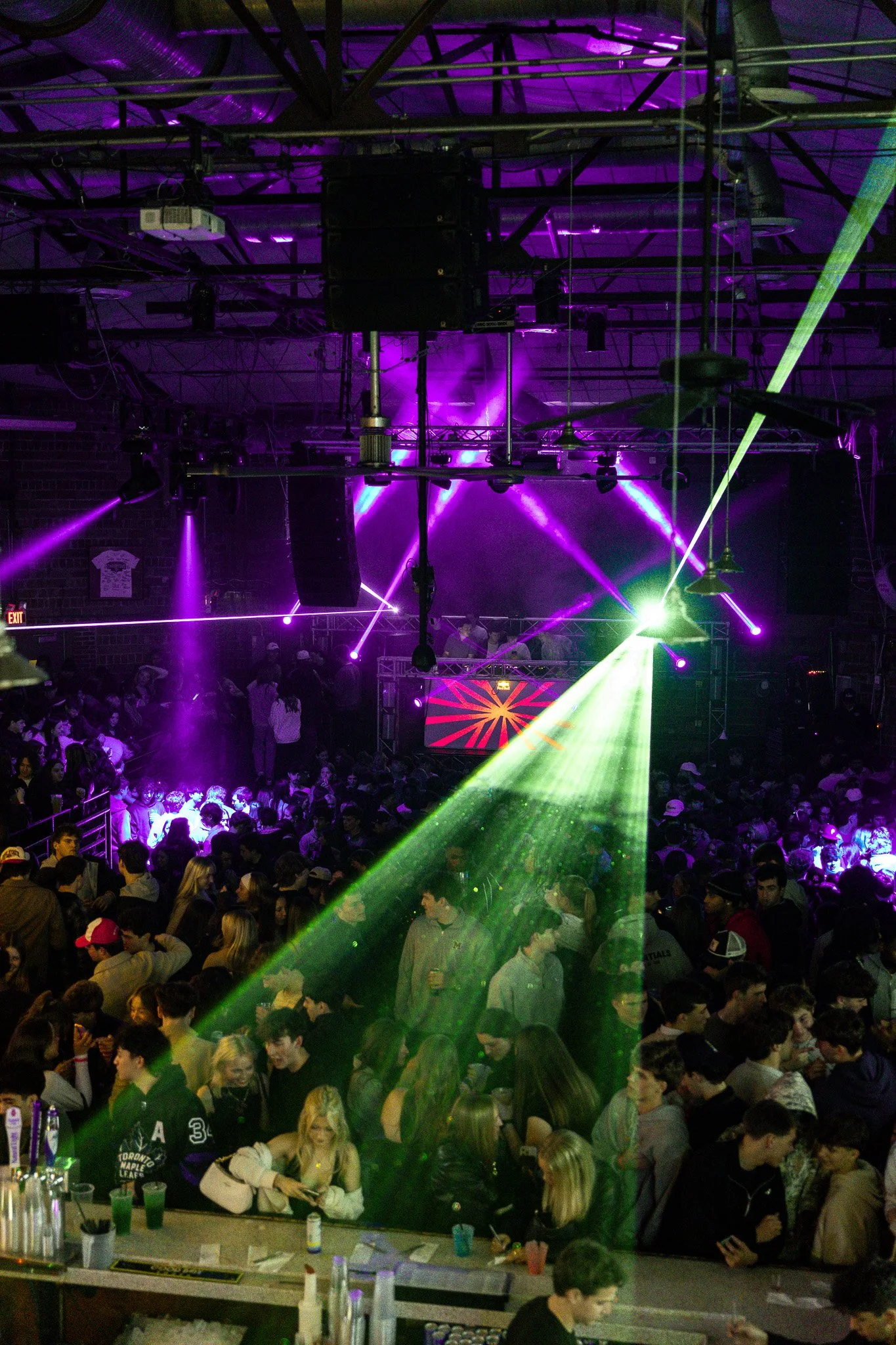 A crowded indoor nightclub with colorful laser lights and a DJ booth on a stage in the background, filled with dancing people.
