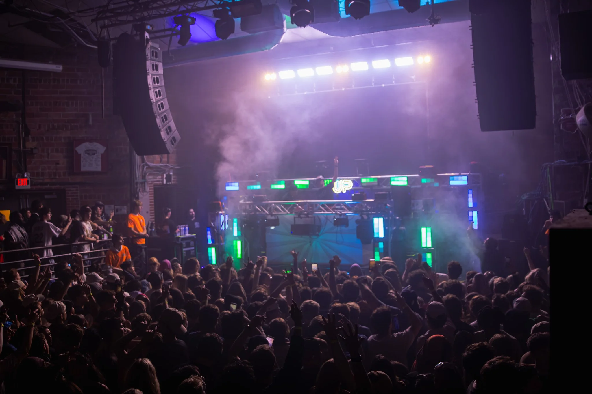 Crowd at a concert or music event with a stage featuring bright LED lights and a DJ or performer in the background, with smoke effects and audience members raising their hands.