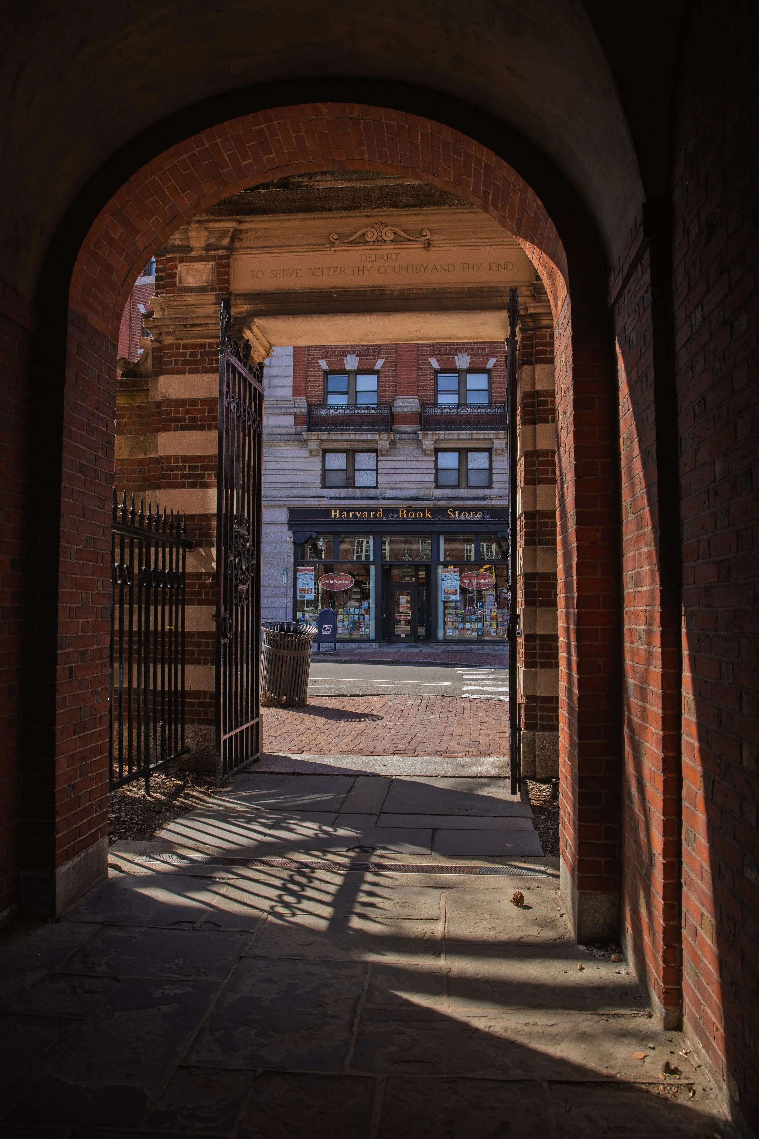View of Harvard Book Store seen through a brick archway with a metal gate. The bookstore is on a city street with posters in the windows and a trash can outside.