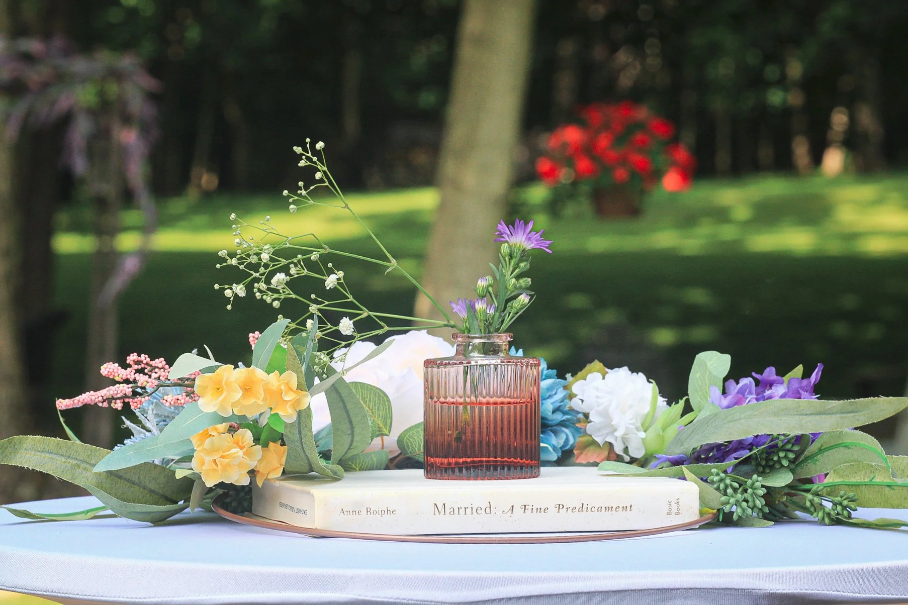 A tray with flowers and a book on a table outdoors. The flowers include yellow, purple, white, and pink blooms, with green leaves. In the center, there is a small pink glass vase with purple flowers. The book titled "Married: A Fine Predicament" by A