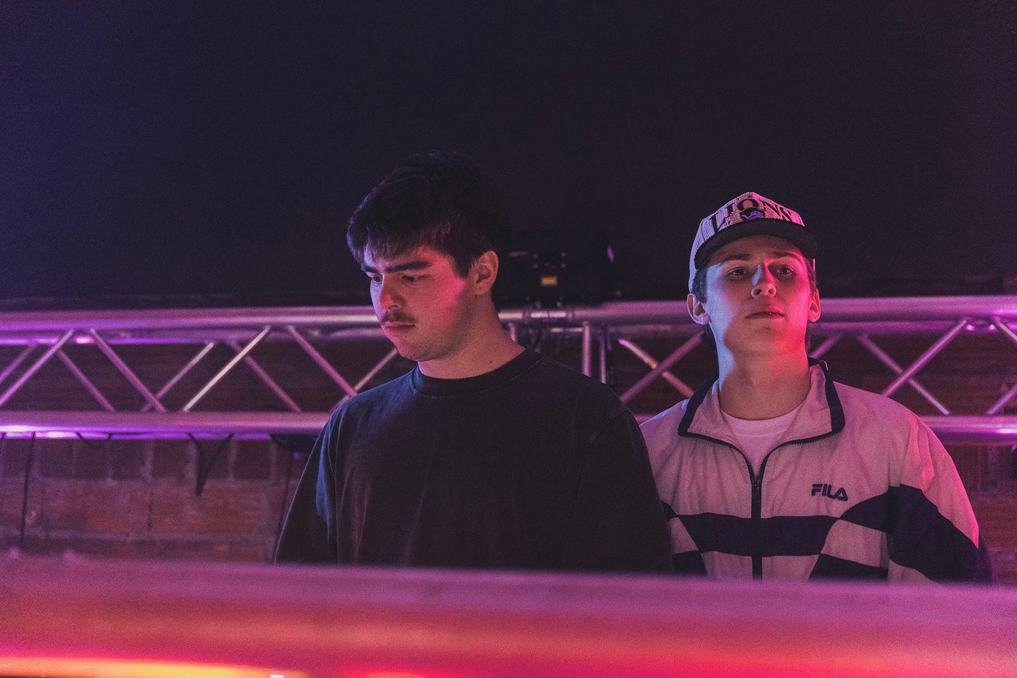 Two young men standing in front of a stage with purple and pink lighting. One wears a black shirt, and the other wears a white FILA jacket and a black cap with 