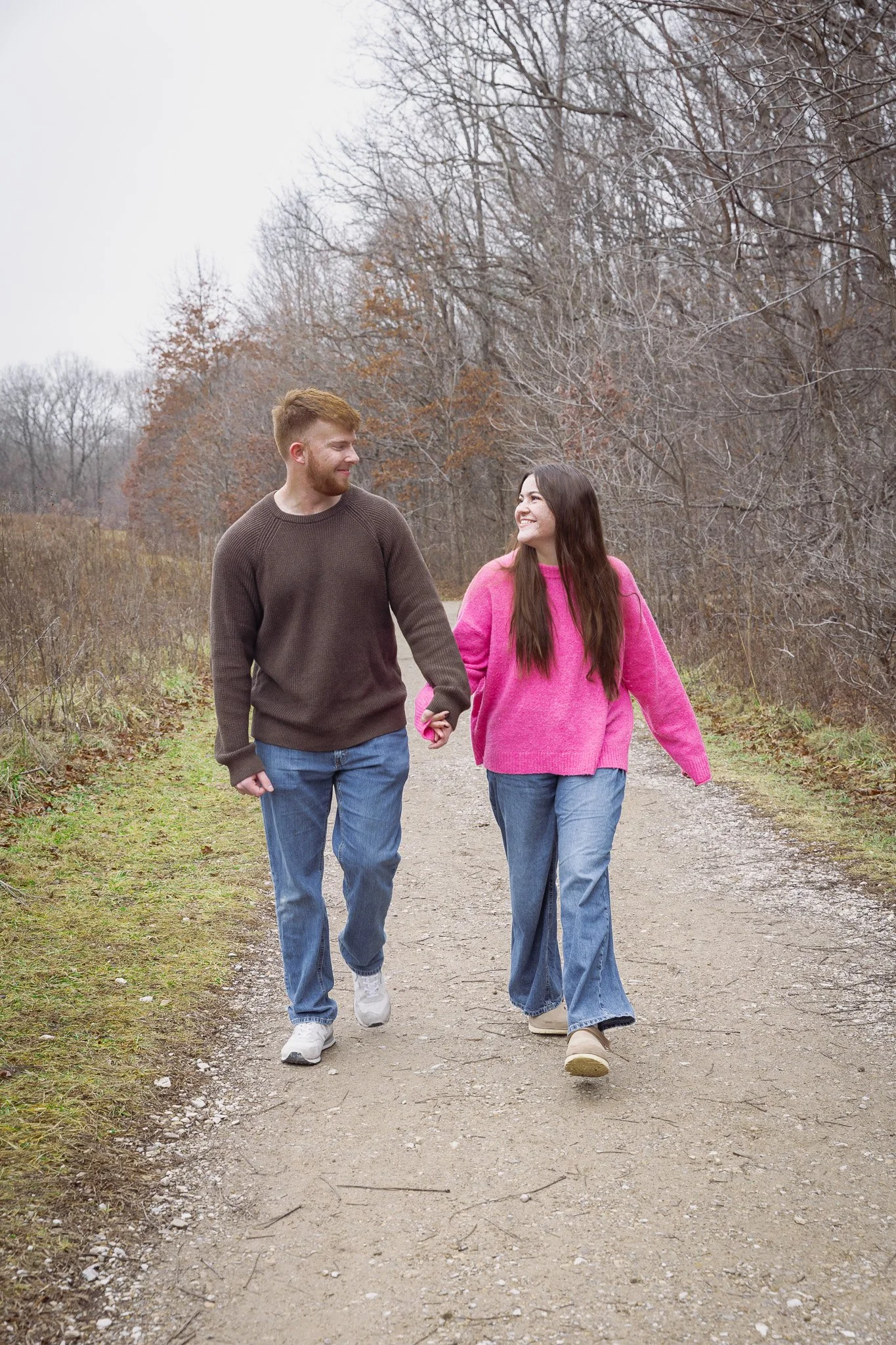 A young man and woman walking hand-in-hand along a dirt path outdoors during fall, with trees in the background.
