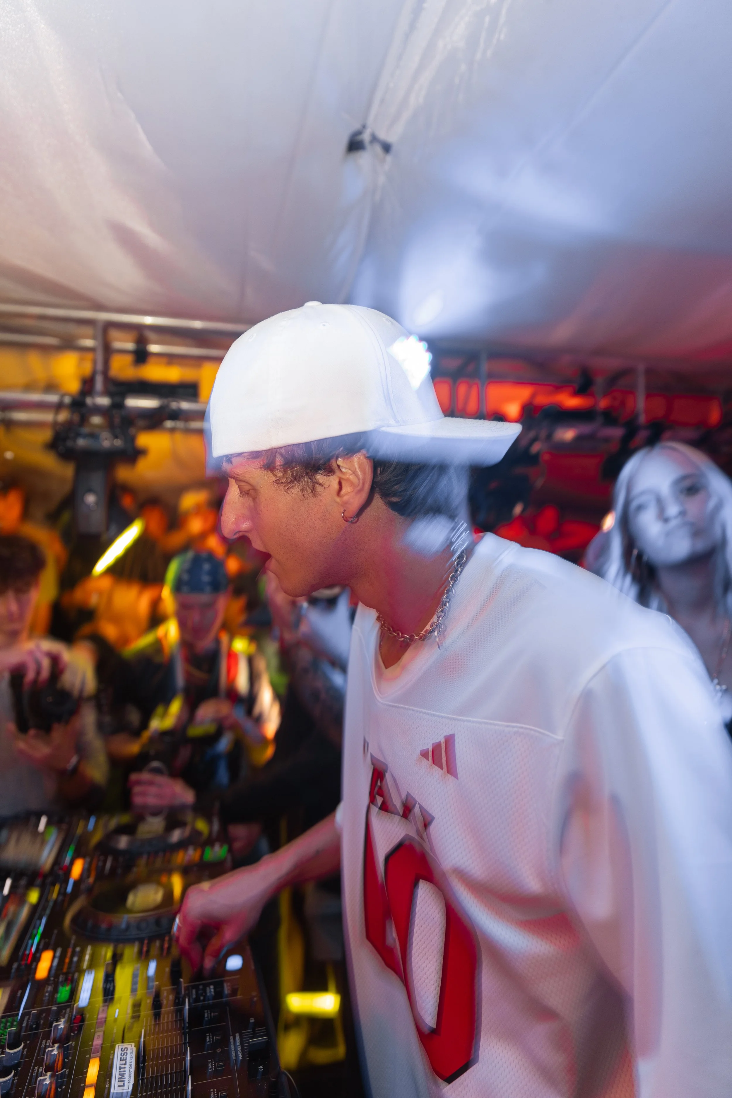 A DJ wearing a white cap and a white sports jersey with red lettering, performing at a crowded indoor event with colorful lighting and people taking photos in the background.