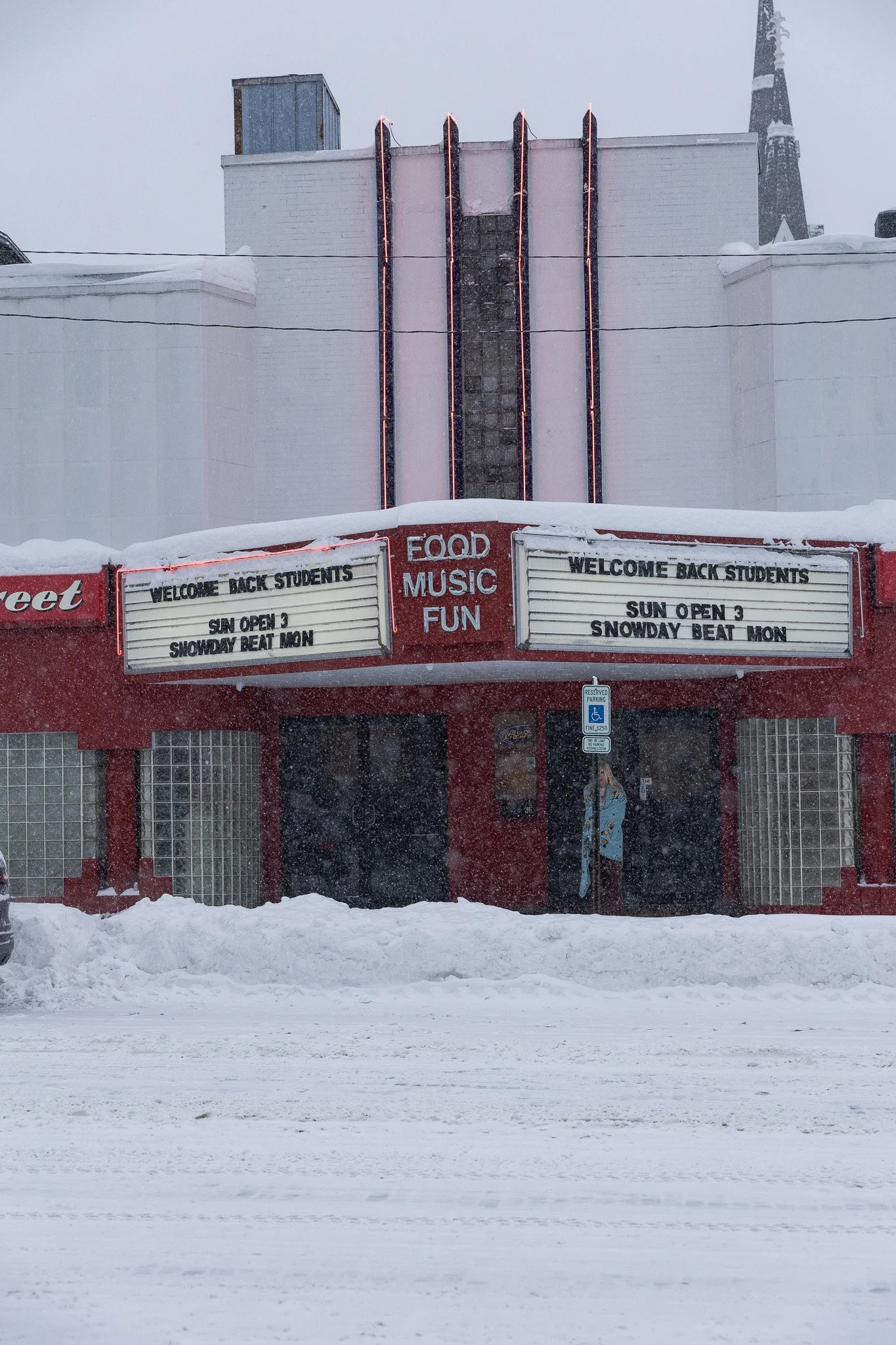 A snow-covered building with a red and white marquee displaying the words "Food, Music, Fun." The marquee also says "Welcome Back Students" and "Sun Open 3, Snowday Beat Mon." There is a person standing in front of the entrance, and a handicapped par