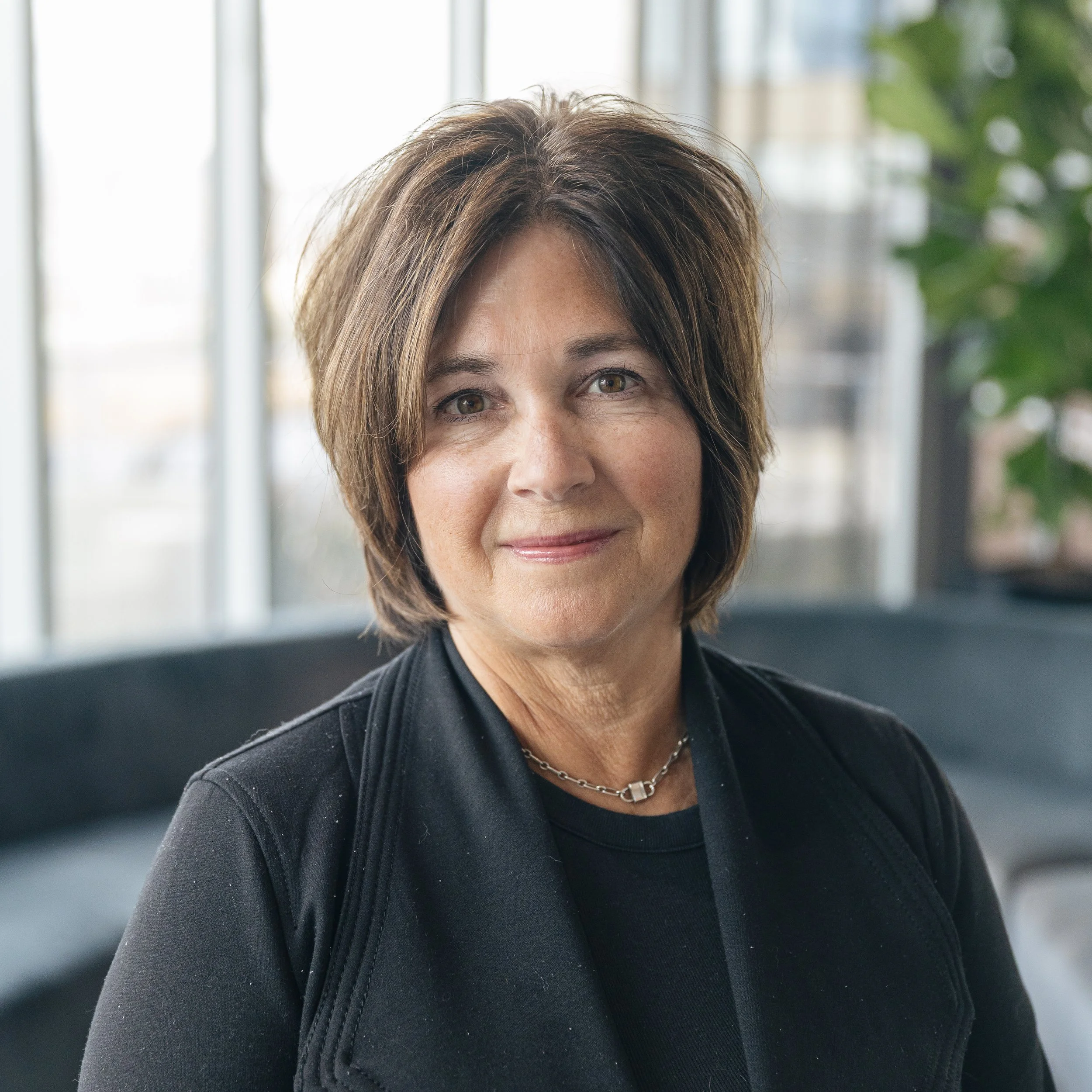 A woman with short brown hair, wearing a black jacket and a silver necklace, smiling at the camera indoors with large windows and a green plant in the background.