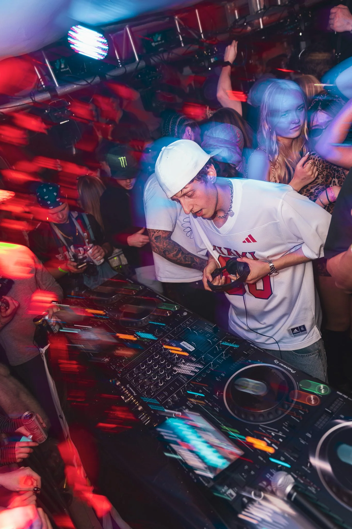 A DJ wearing a white cap and Miami Hurricanes jersey mixes music at a crowded nightclub with colorful lights and dancing people around him.