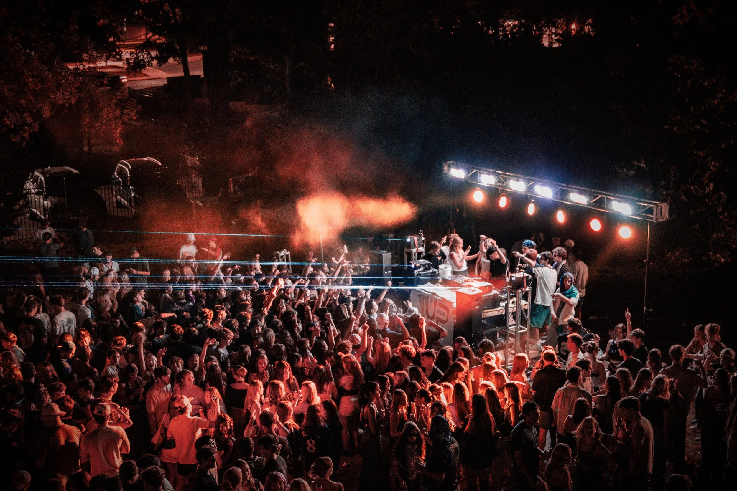 A large crowd of people dancing and enjoying a live outdoor concert at night. The stage is lit with bright lights and there is smoke or fog in the air.