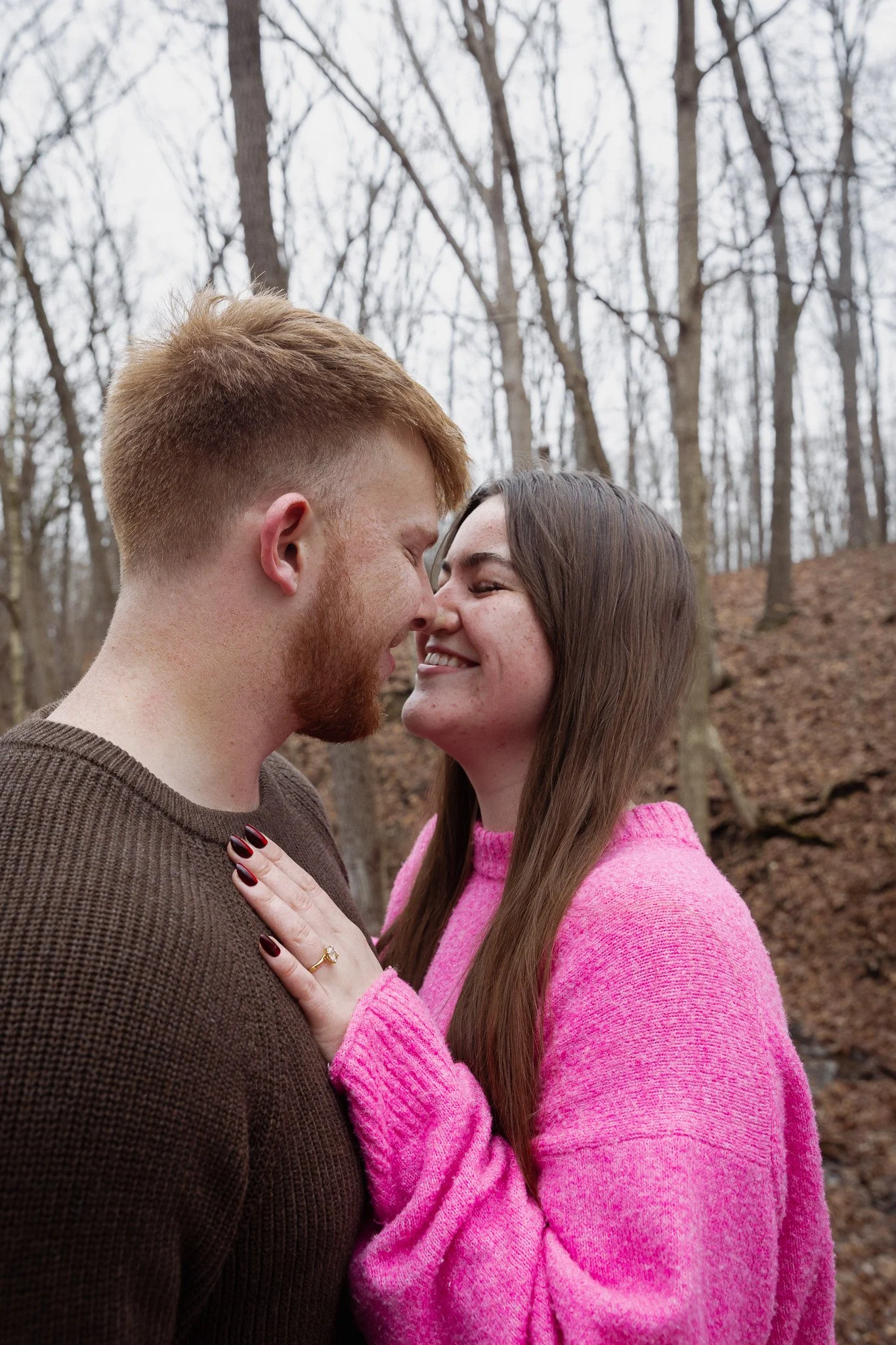 A couple standing close together outdoors in a wooded area, smiling and touching foreheads.
