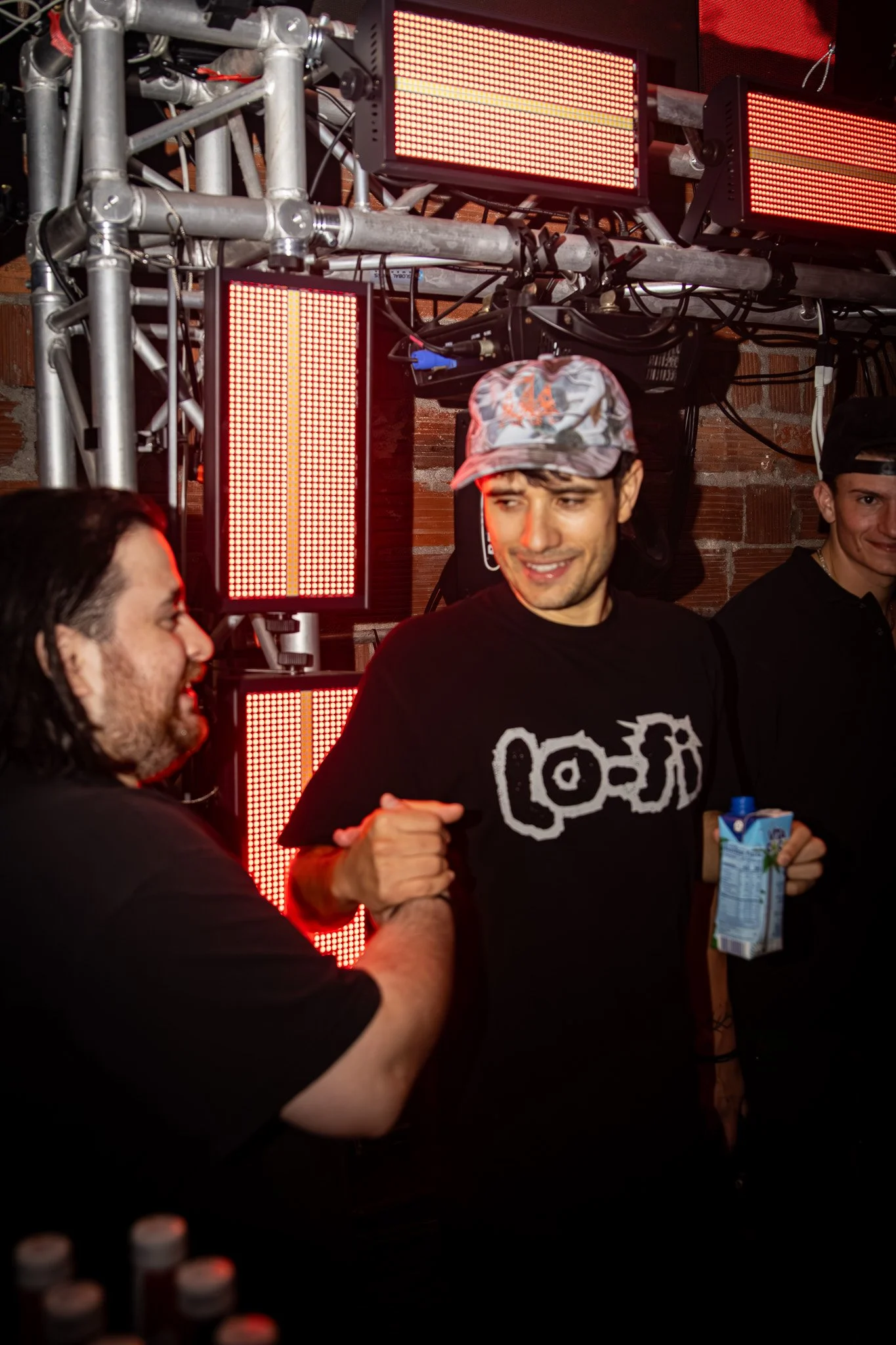 Two men handshake, one wearing a black t-shirt, the other with a cap, surrounded by red LED lights in an indoor setting.