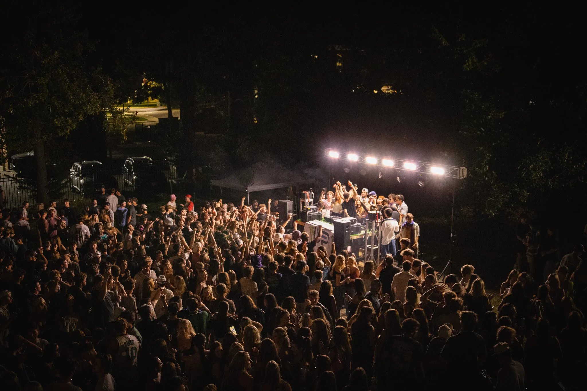 A large crowd of people dancing and enjoying music at an outdoor night concert with bright stage lights.