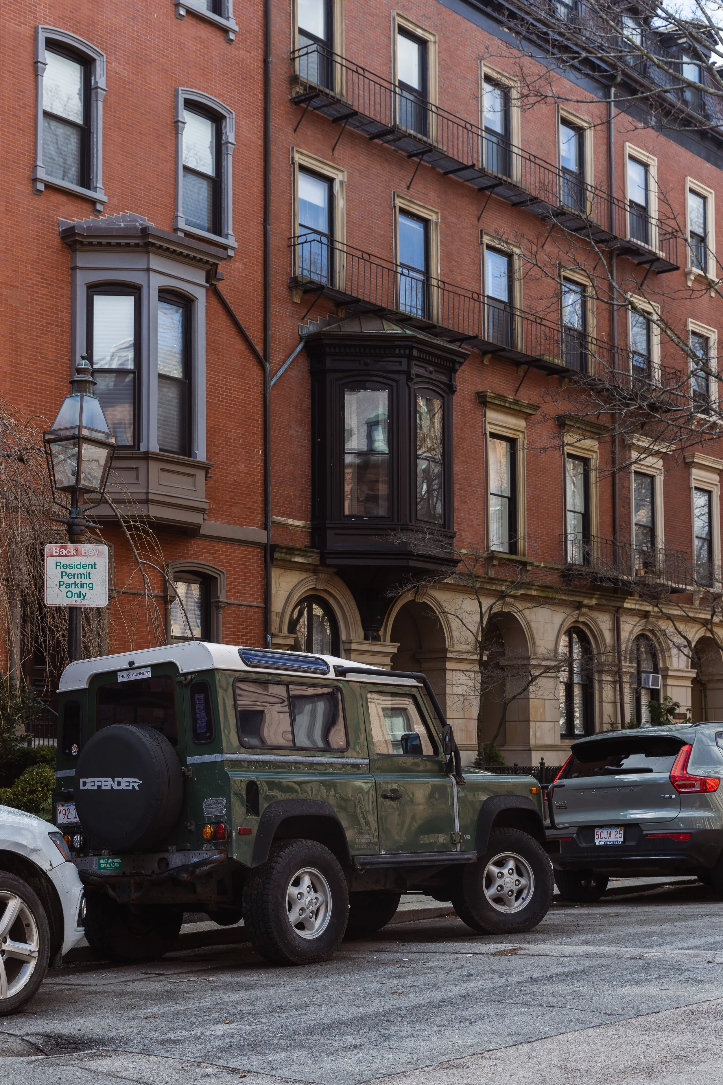 A multi-story residential brick building with bay windows and black metal fire escape balconies on the front. A green Land Rover Defender is parked in front, along with other cars, on a city street.
