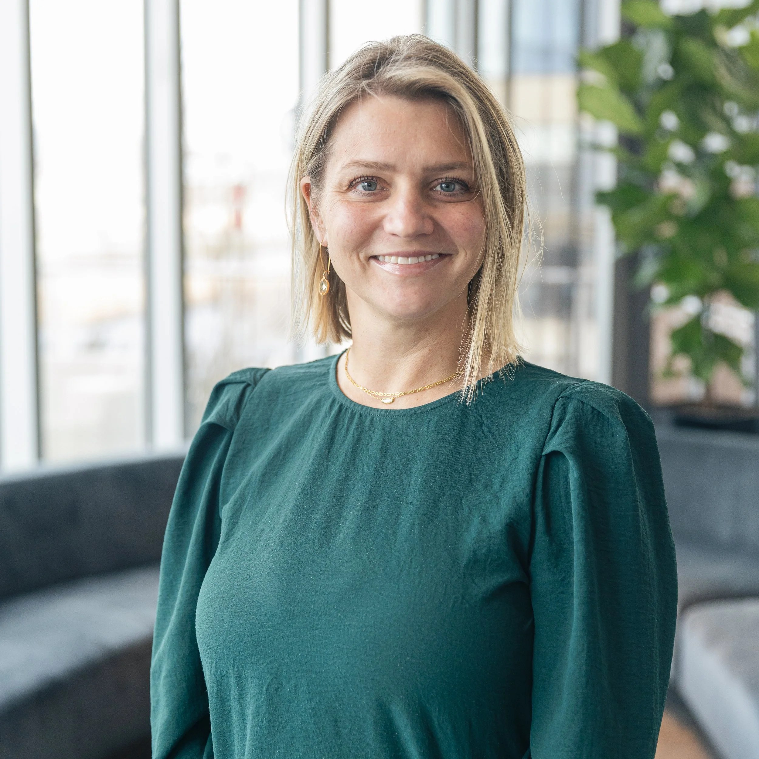 A woman with blonde hair and blue eyes is smiling while standing indoors near a large window with a plant in the background.