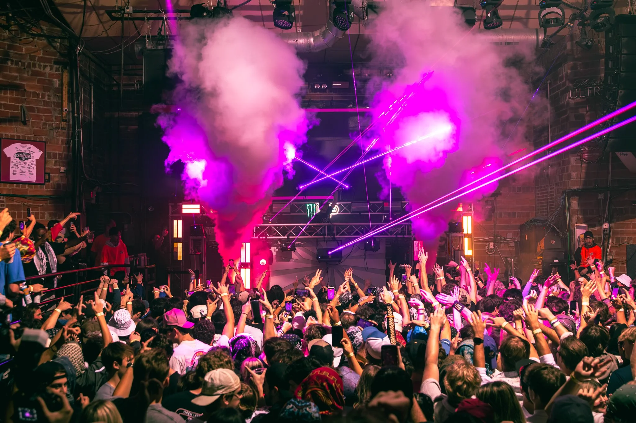 A crowded indoor concert with a DJ on stage, colorful lights, smoke effects, and laser beams, with people dancing and taking photos.