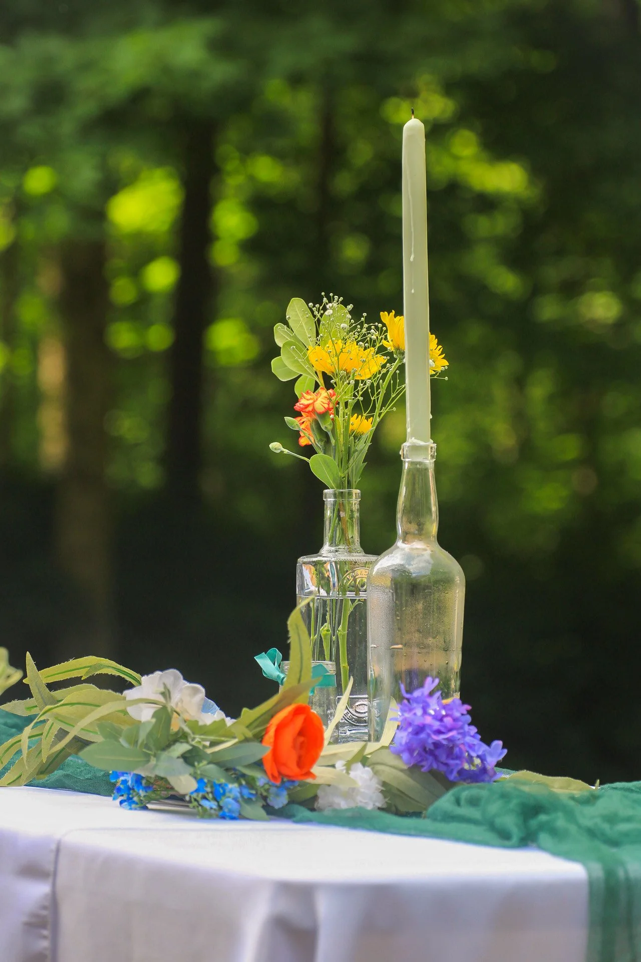 A table with a green cloth runner, decorated with colorful flowers and a white candle in a tall glass holder, set outdoors with a blurred green forest background.