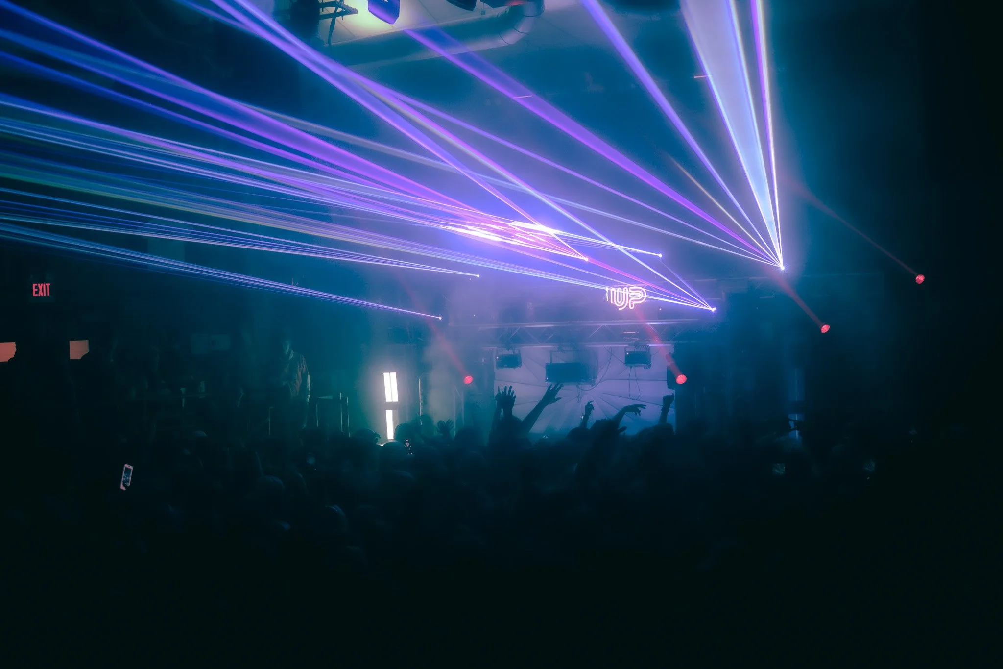 A dark concert venue filled with vibrant purple and blue laser lights, a crowd of people dancing and raising their hands, with a DJ booth and illuminated sign in the background.